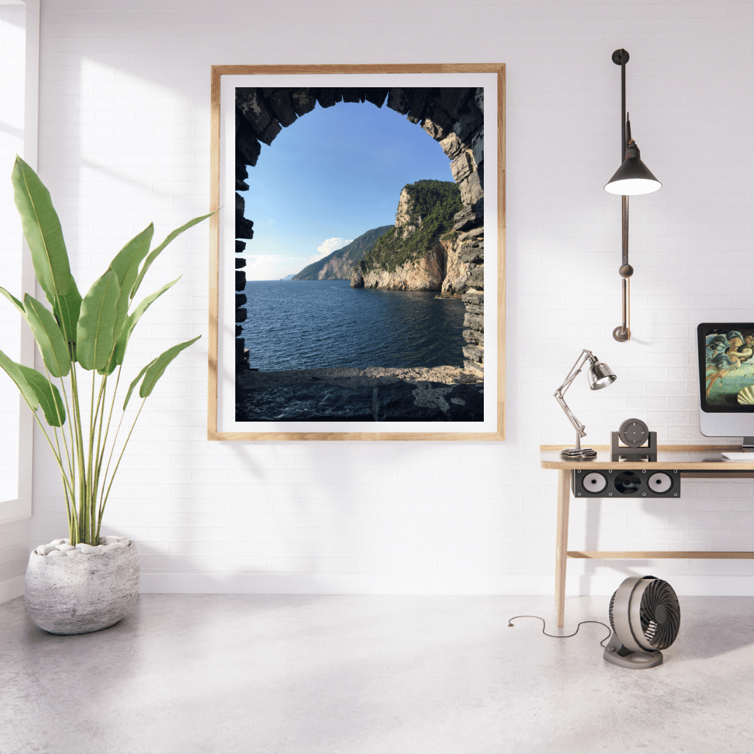 A framed vertical art print depicting a scenic coastal view from a stone window, displayed on a white wall in a modern, sunlit room with a potted plant, desk, lamp, and computer.