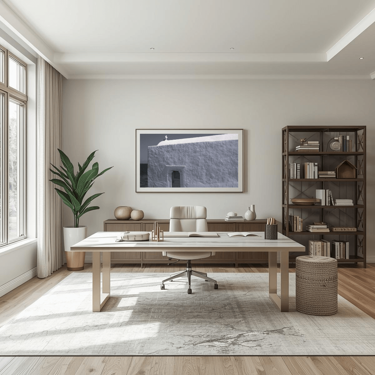 A modern home office interior featuring a light wood writing desk as the central furniture piece, styled with a white office chair, a large area rug, a potted plant, a dark wood bookshelf, and a framed black and white print on the wall depicting a small cross on top of a whitewashed building in Mykonos, Greece.