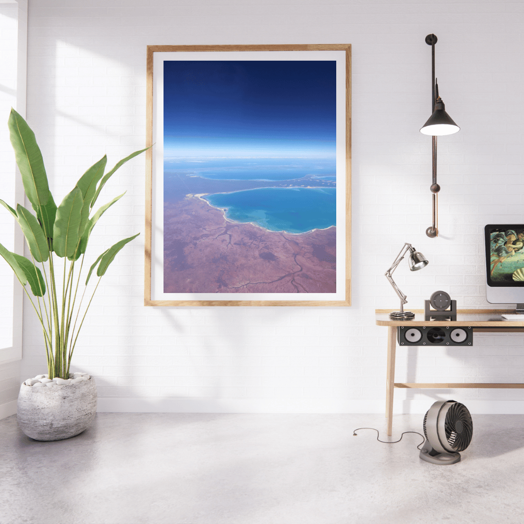 A framed aerial landscape photograph of an aerial view of the deeply contrasting deep blue ocean against the red earth along the Western Australian coastline, displayed on a white wall in a modern room setting with a plant, desk, lamp, and computer.