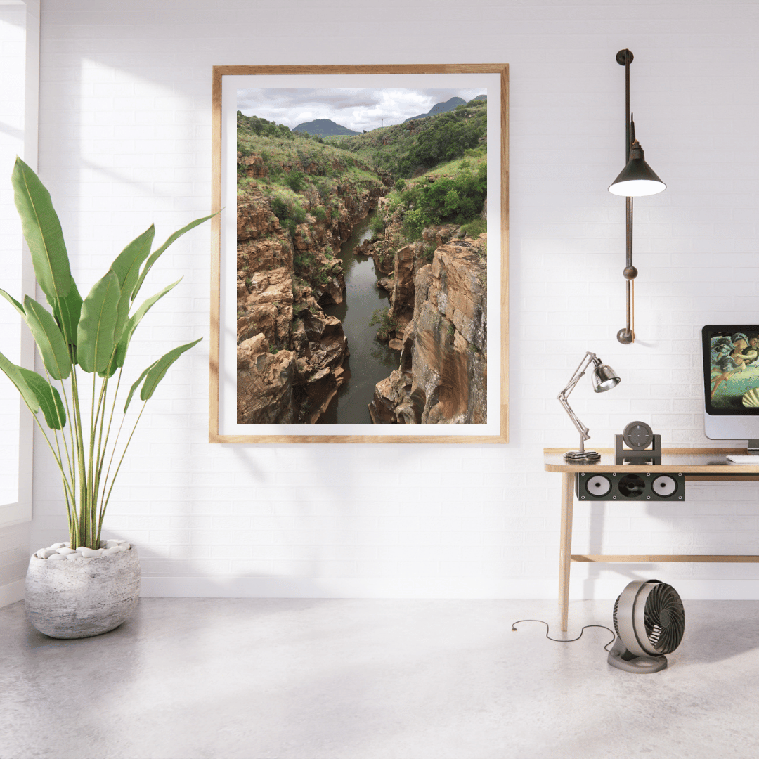 A framed landscape art print of a deep rocky canyon with a river flowing through it, displayed on a white wall in a modern, sunlit room with a potted plant, desk, and lamp.