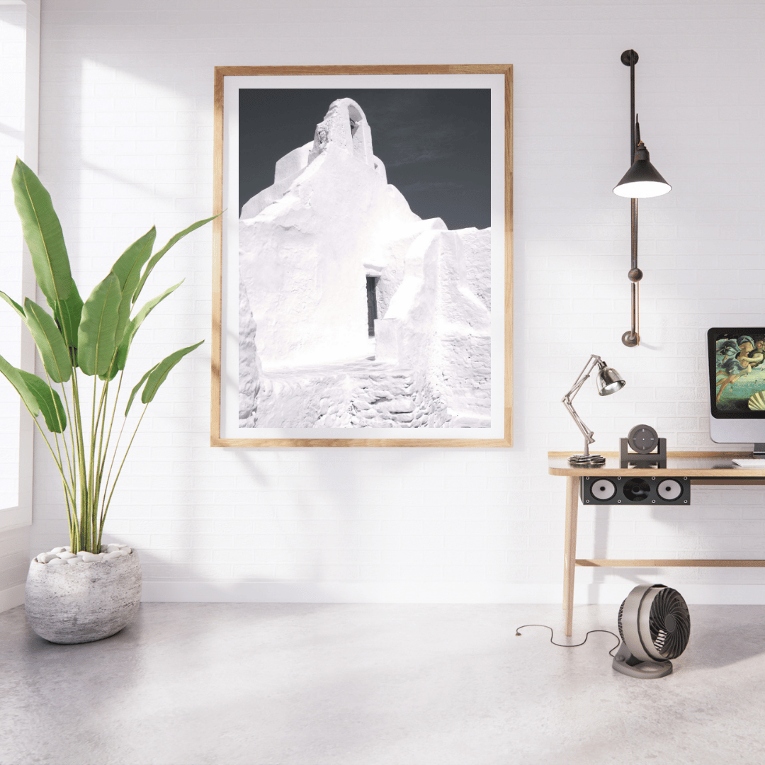 A framed black and white art print featuring a whitewashed building in Mykonos, Greece hangs on a white wall in a modern, sunlit home office. The room features a wooden desk with a computer, a potted green plant, and a floor lamp.