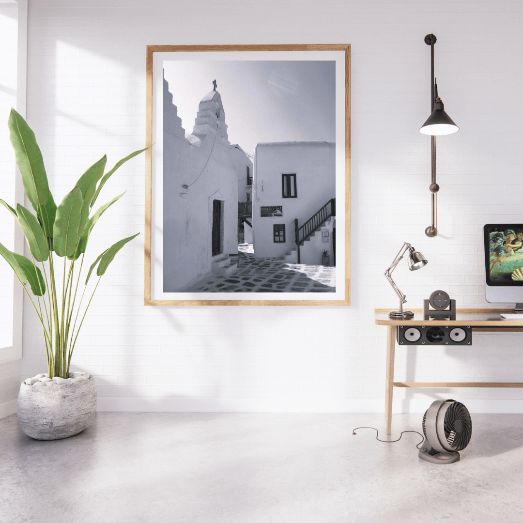 A framed black and white architectural photograph of white buildings and a staircase, displayed on a white wall in a modern room setting with a plant, desk, lamp, and computer.