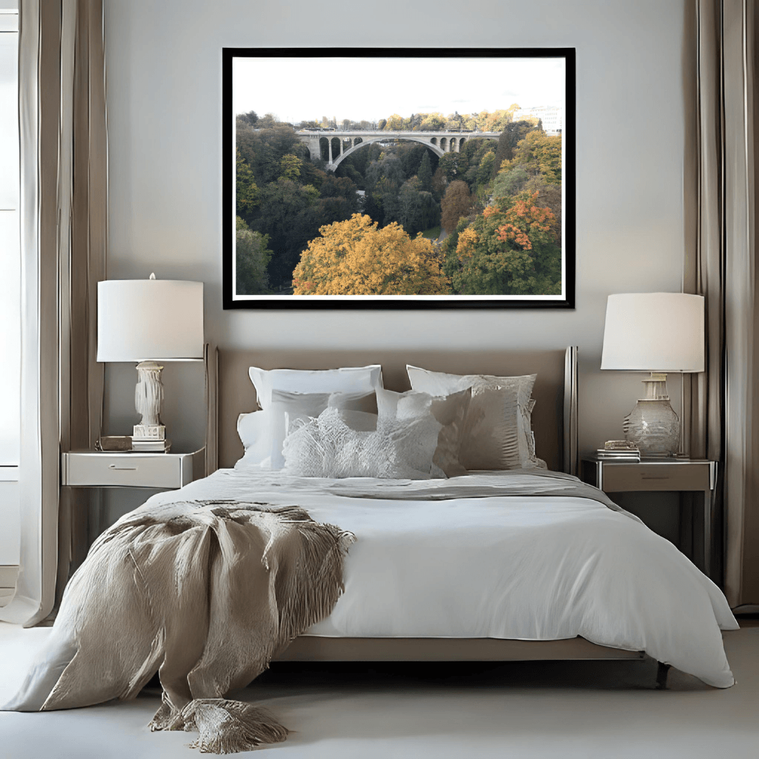 A framed landscape art print depicting Luxembourg's Adolphe bridge over a forest in autumn, displayed on a neutral gray wall above a modern bed in a styled bedroom.