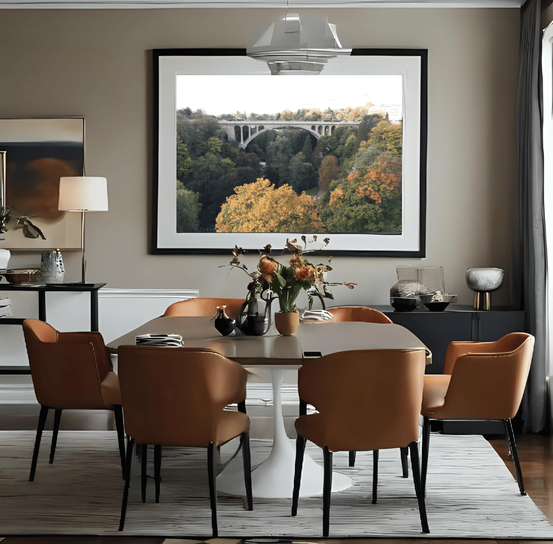 A large framed art print depicting a scenic landscape with Luxembourg's Adolphe bridge over autumn-colored trees, displayed on a beige wall in a modern dining room setting with brown leather chairs and a round dining table.
