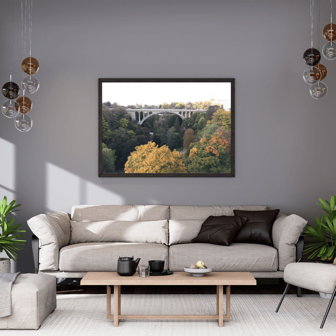 A framed landscape art print of Luxembourg's Adolphe bridge spanning a forest canyon in autumn, displayed on a gray wall above a modern beige sofa in a styled living room.