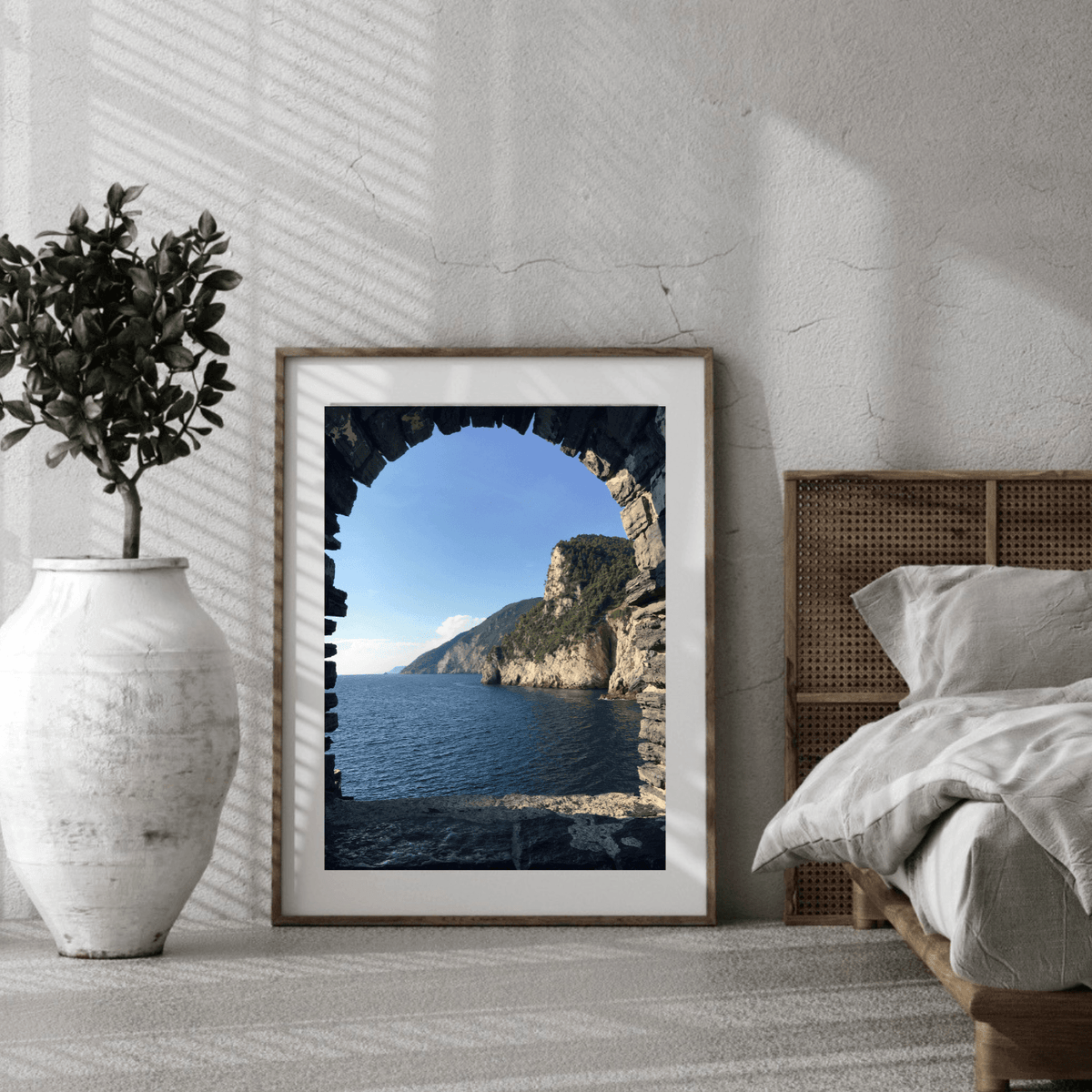A framed coastal landscape print leaning against a textured white wall in a styled bedroom. The artwork shows a stone archway framing a blue sea and green cliffside landscape.
