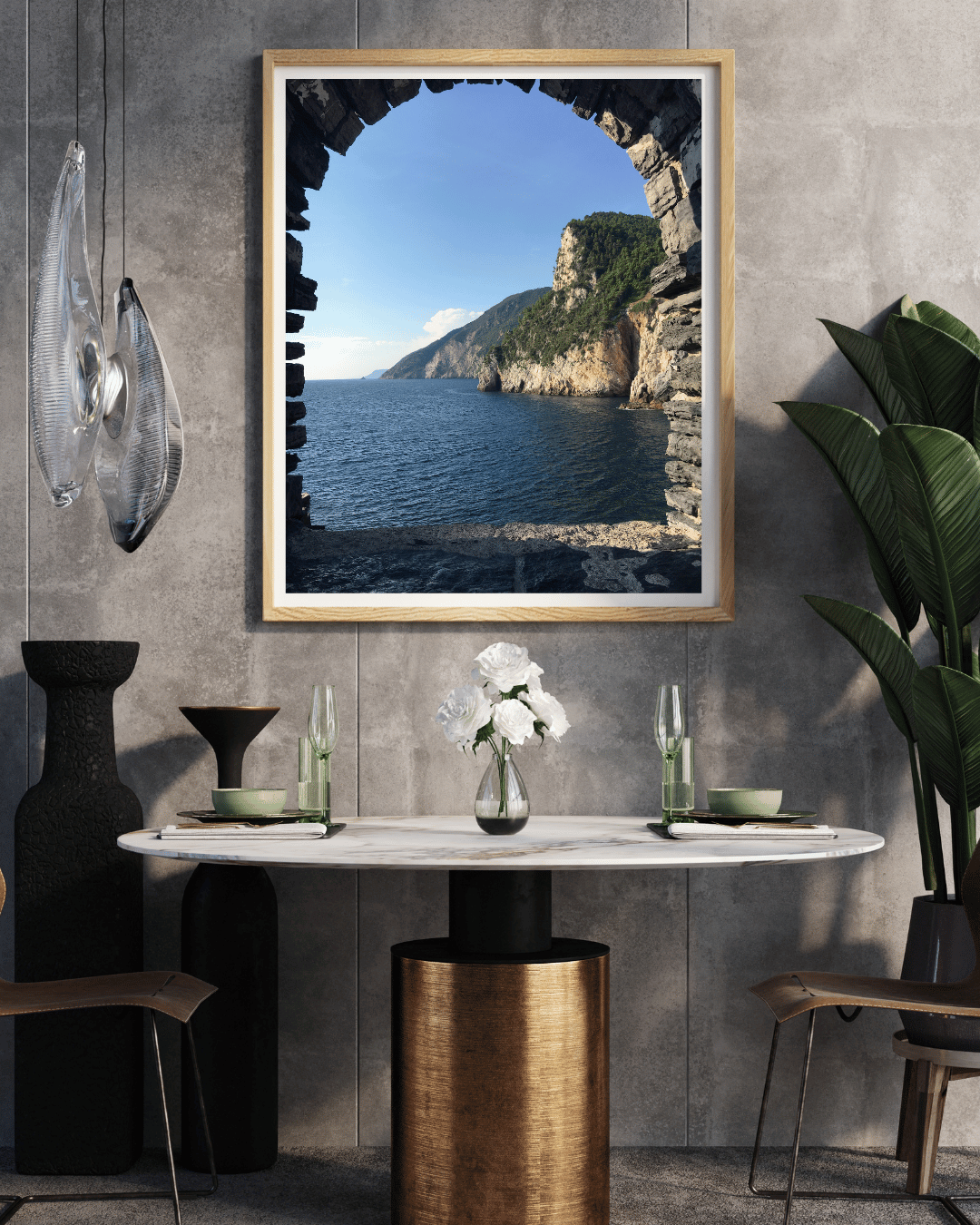 A framed art print depicting a scenic coastal landscape seen through a stone archway, displayed on a textured gray wall above a dining table in a modern styled room.