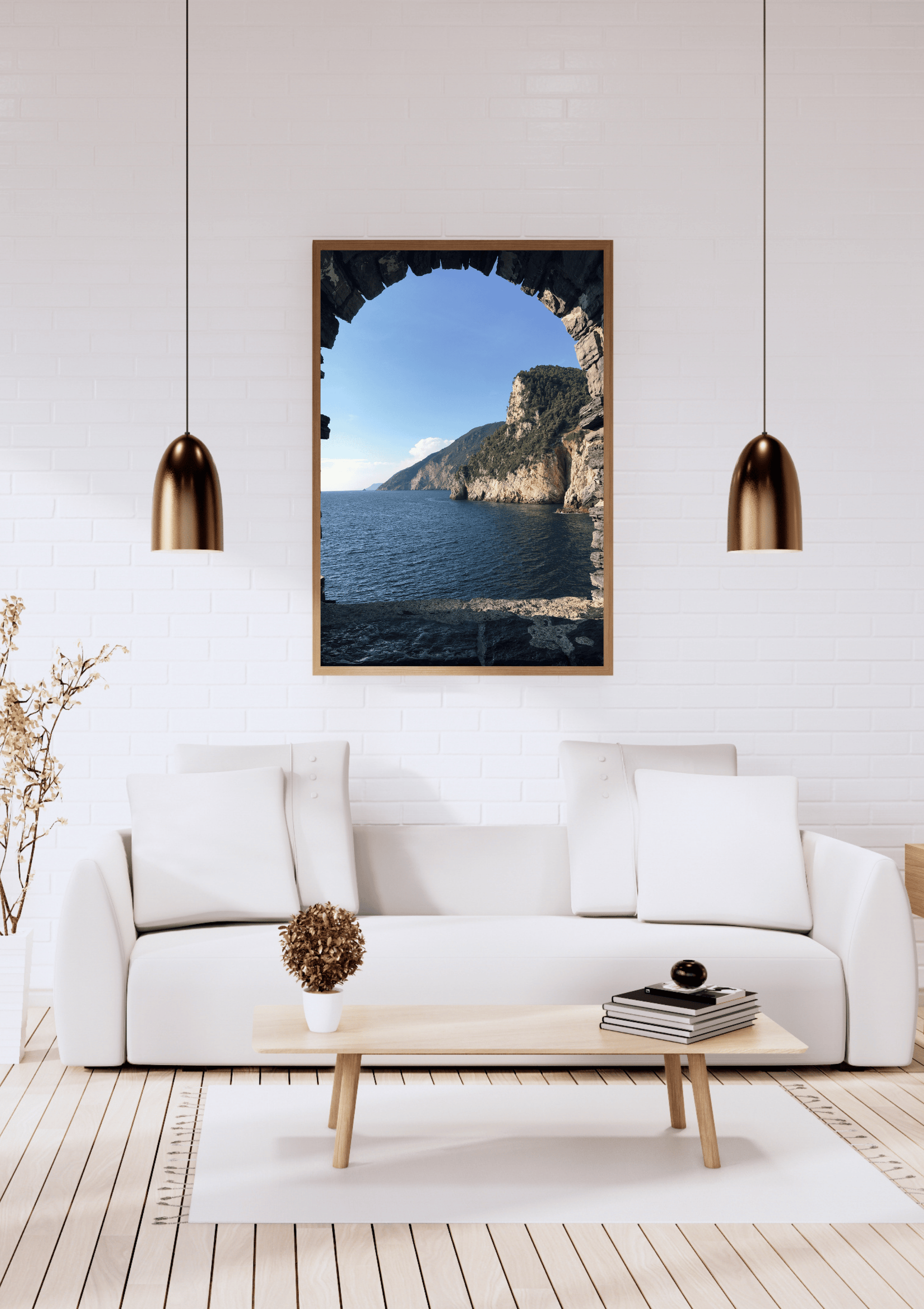 A framed art print depicting a coastal landscape seen through a stone archway, displayed on a white brick wall above a modern white sofa in a bright, contemporary living room.