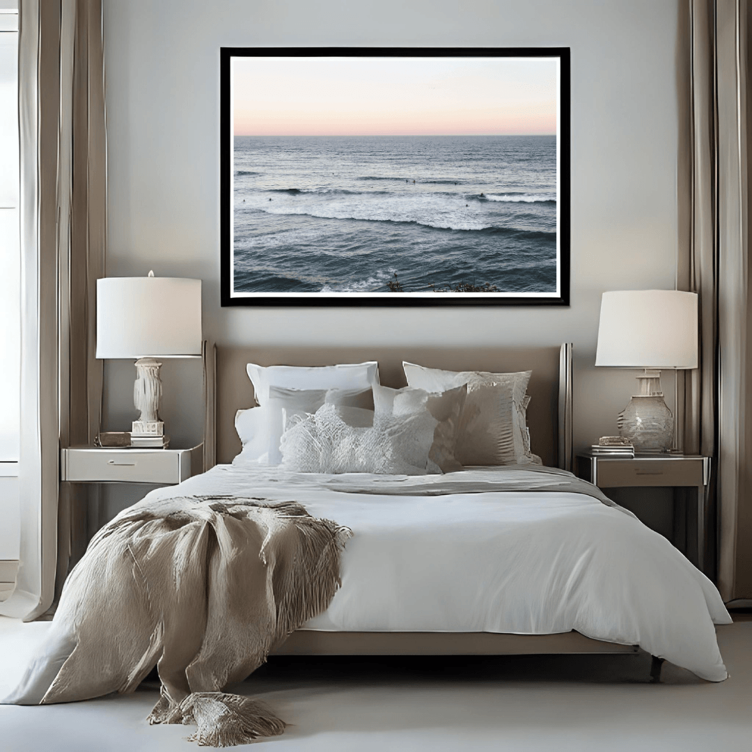 A framed horizontal ocean sunset print hangs above a neatly made bed in a modern, symmetrically styled bedroom with beige curtains, white bedding, and matching bedside tables and lamps.