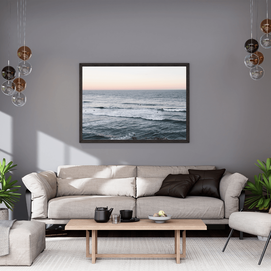 A framed horizontal ocean landscape print featuring the iconic Bondi Beach with a few swimmers capturing the soft light of sunset, hangs on a gray wall above a light beige sofa in a modern living room setting.