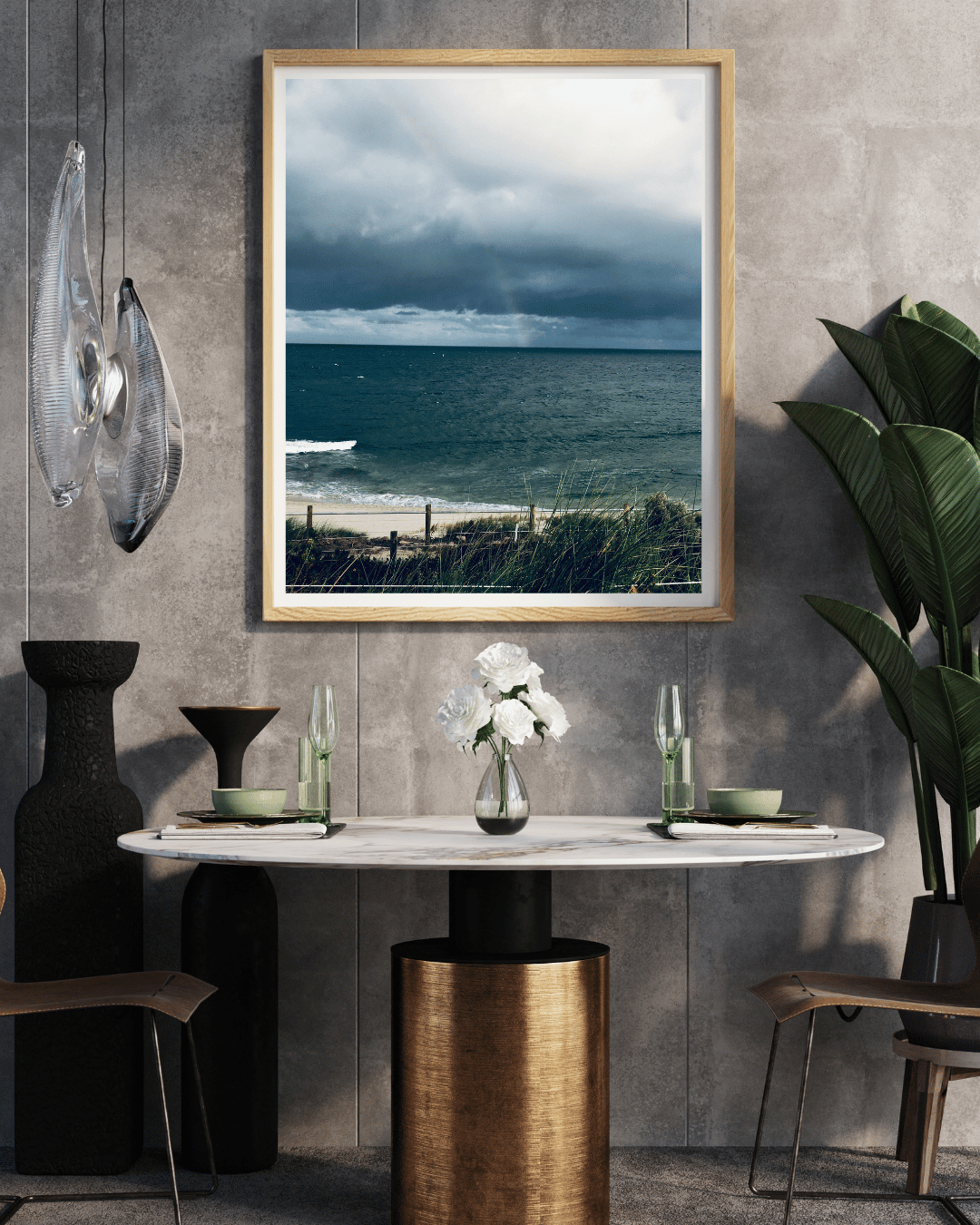A framed art print depicting the darker moody colours of the sky and beach before a storm in Cottesloe, Western Australia, above a modern dining table with plants and decorative vases.