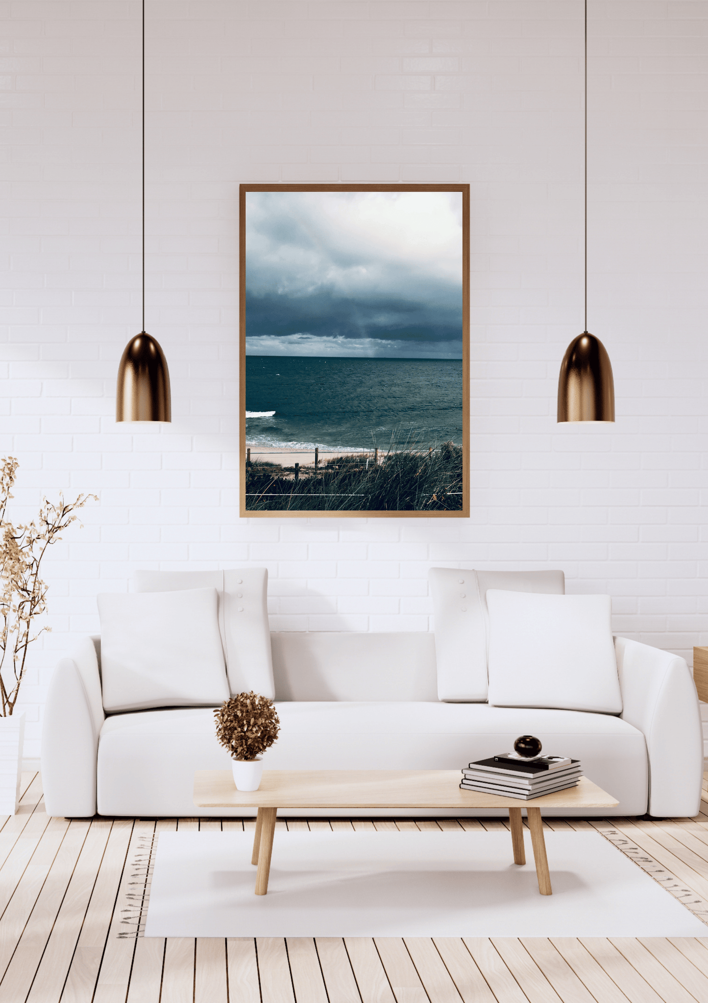 A framed vertical landscape print of a featuring the darker moody colours of the sky and beach before a storm in Cottesloe, Western Australia hangs on a white brick wall above a modern white sofa in a bright living room. 