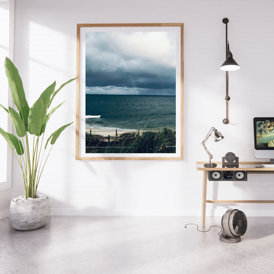 A framed vertical art print of the darker moody colours of the sky and beach before a storm in Cottesloe, Western Australia hangs on a white wall in a modern, sunlit room with a potted plant, desk, lamp, and computer monitor.