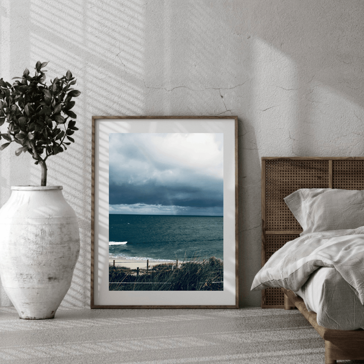 A framed coastal landscape print featuring the darker moody colours of the sky and beach before a storm in Cottesloe, Western Australia, leaning against a textured white wall in a styled bedroom, next to a white vase with a potted plant and a bed with white bedding.