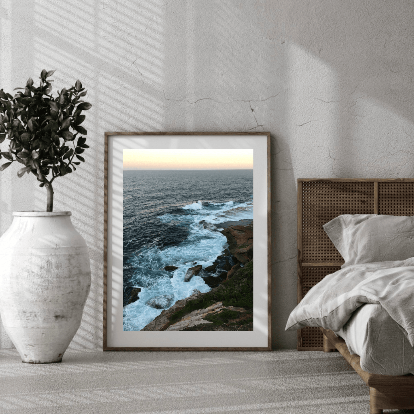 A framed coastal landscape art print leaning against a textured wall in a modern bedroom setting, next to a bed and a large white vase with a plant.