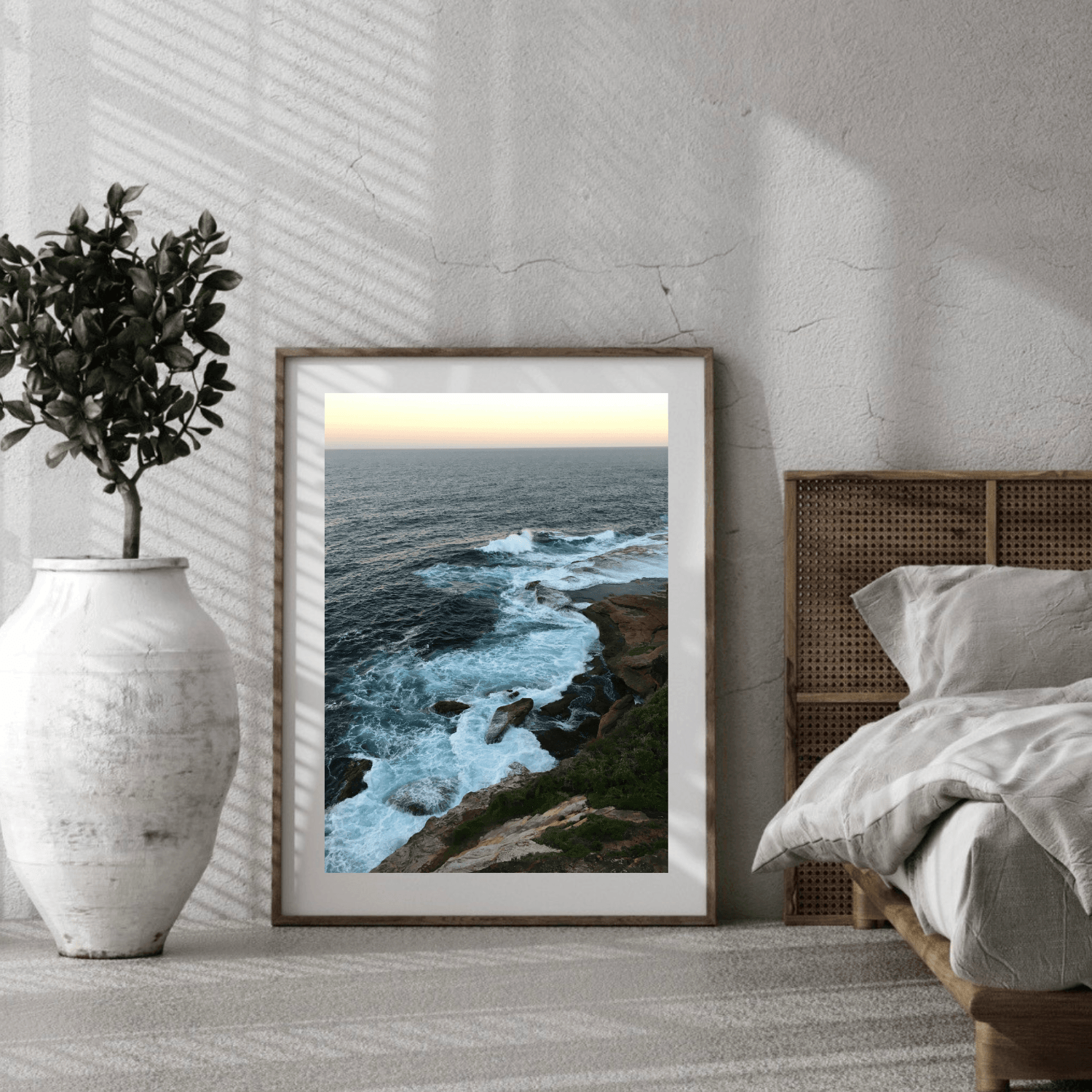 A framed coastal landscape art print leaning against a textured wall in a modern bedroom setting, next to a bed and a large white vase with a plant.
