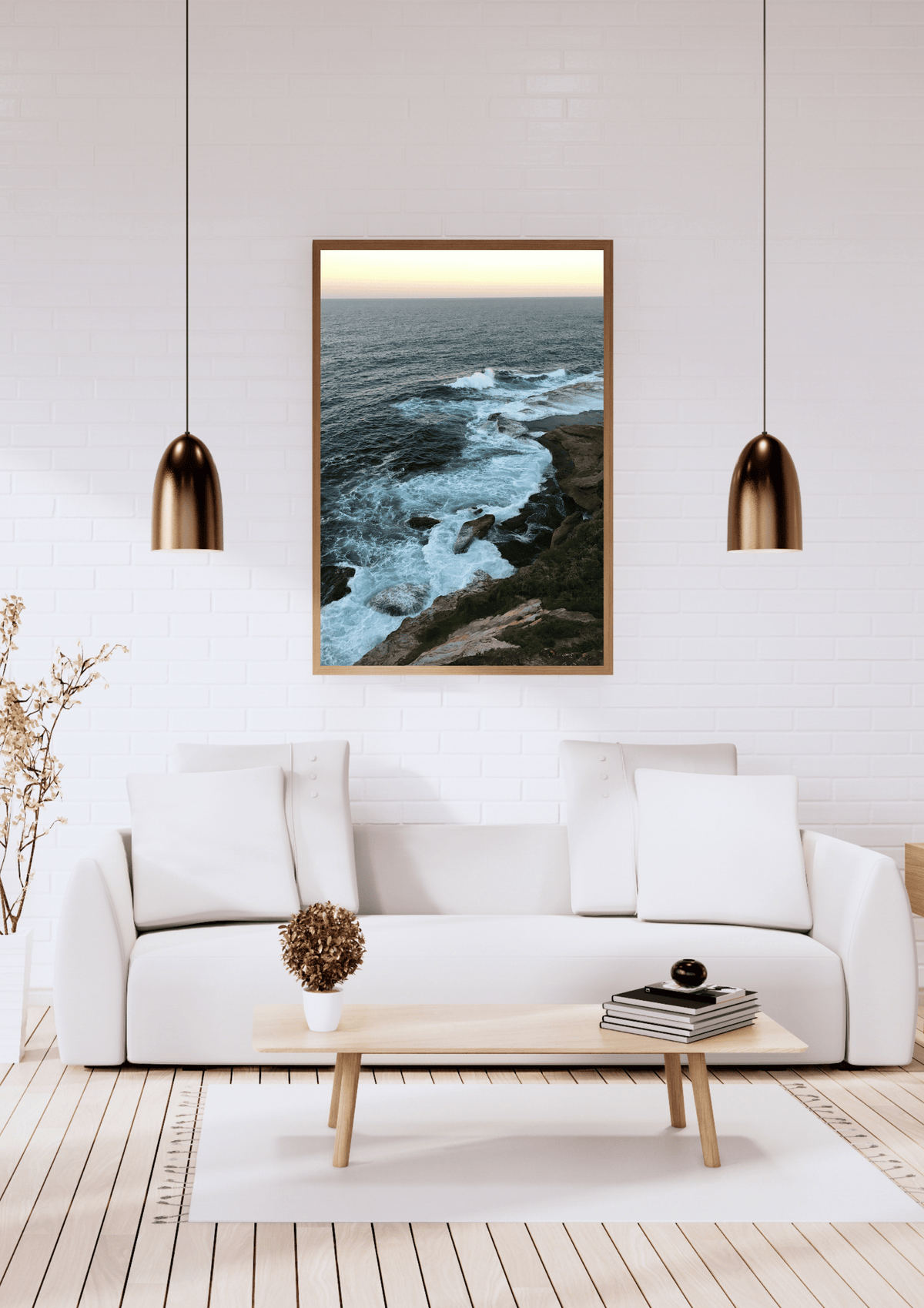A framed vertical landscape print of a rocky coastline with crashing waves, hanging on a white brick wall above a modern white sofa in a bright living room.