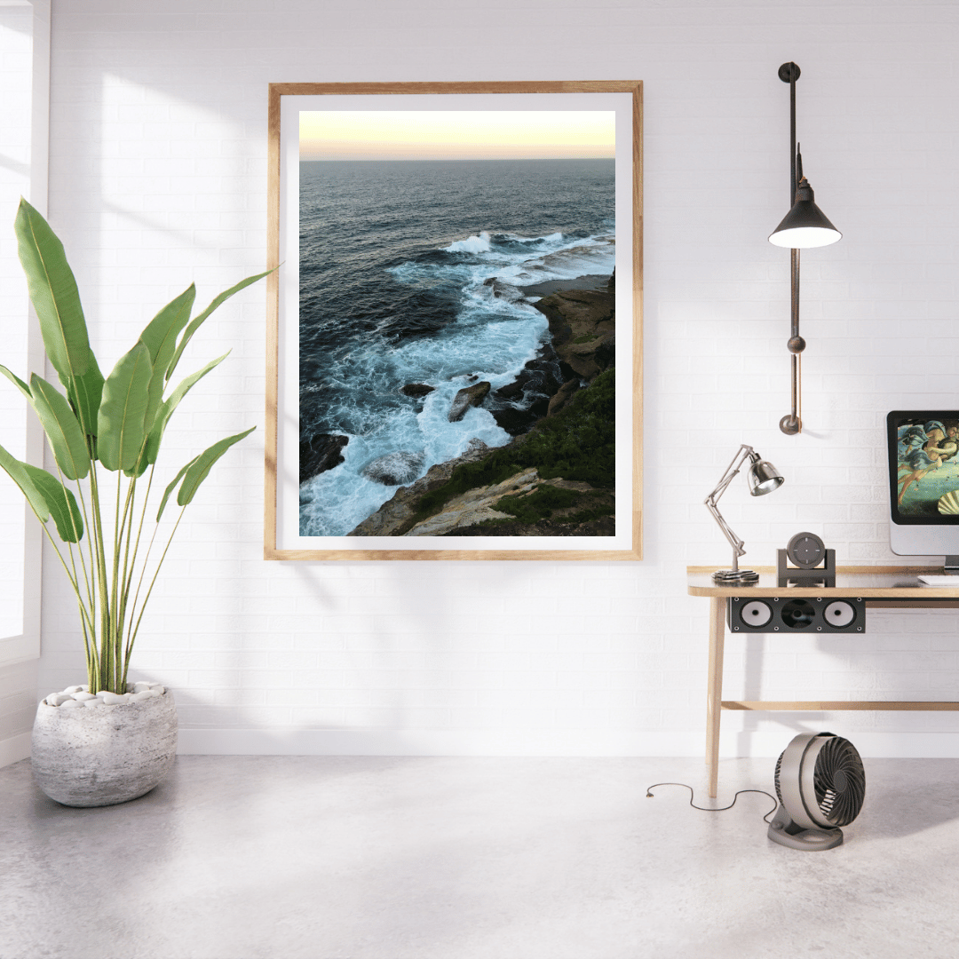 A framed vertical art print of a rocky coastline with ocean waves is displayed on a white wall in a modern, sunlit room with a potted plant, desk, lamp, and computer.