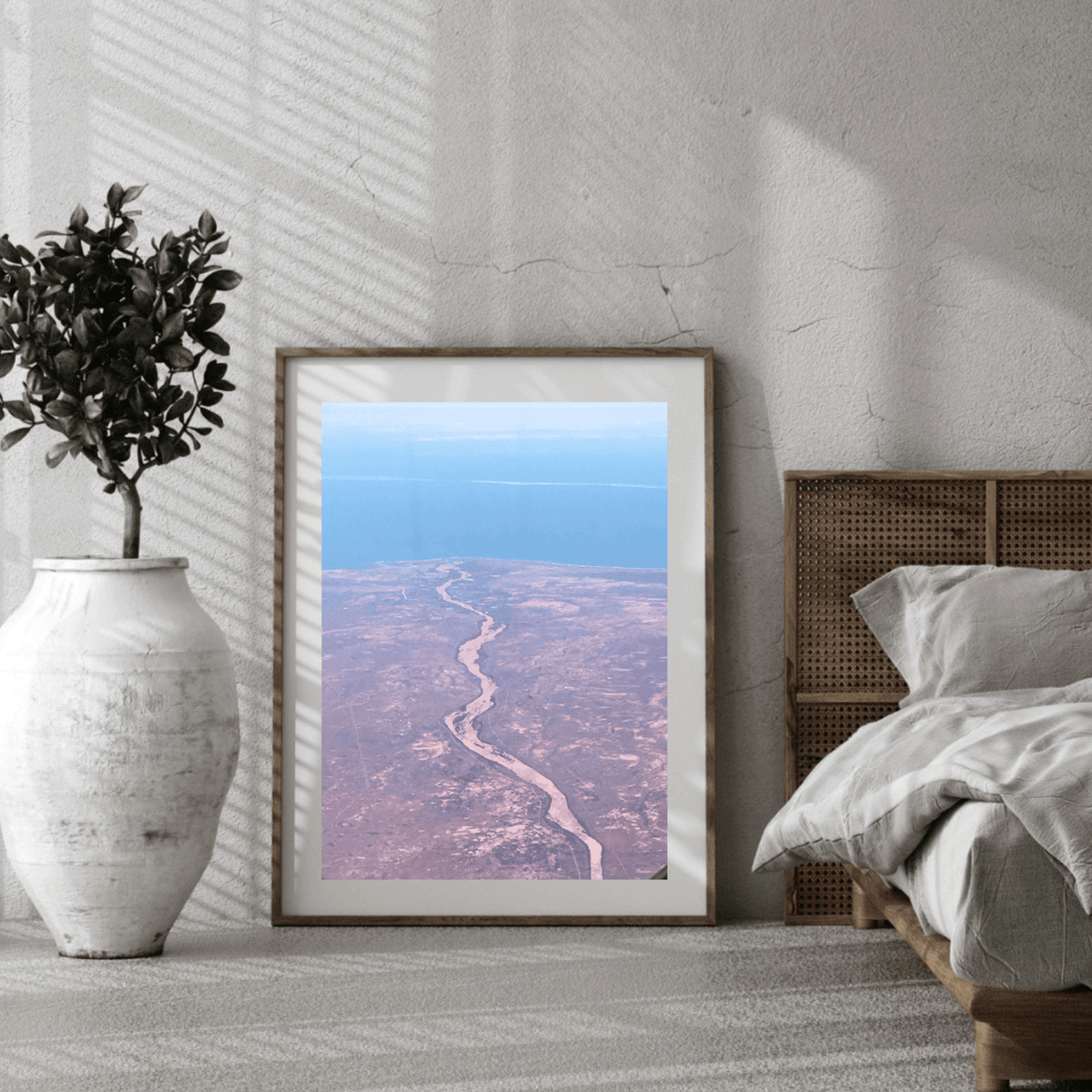 A framed art print depicting an aerial view of the crimson colour of the barren land in Australia, styled in a modern bedroom setting next to a bed and a white vase with a plant.