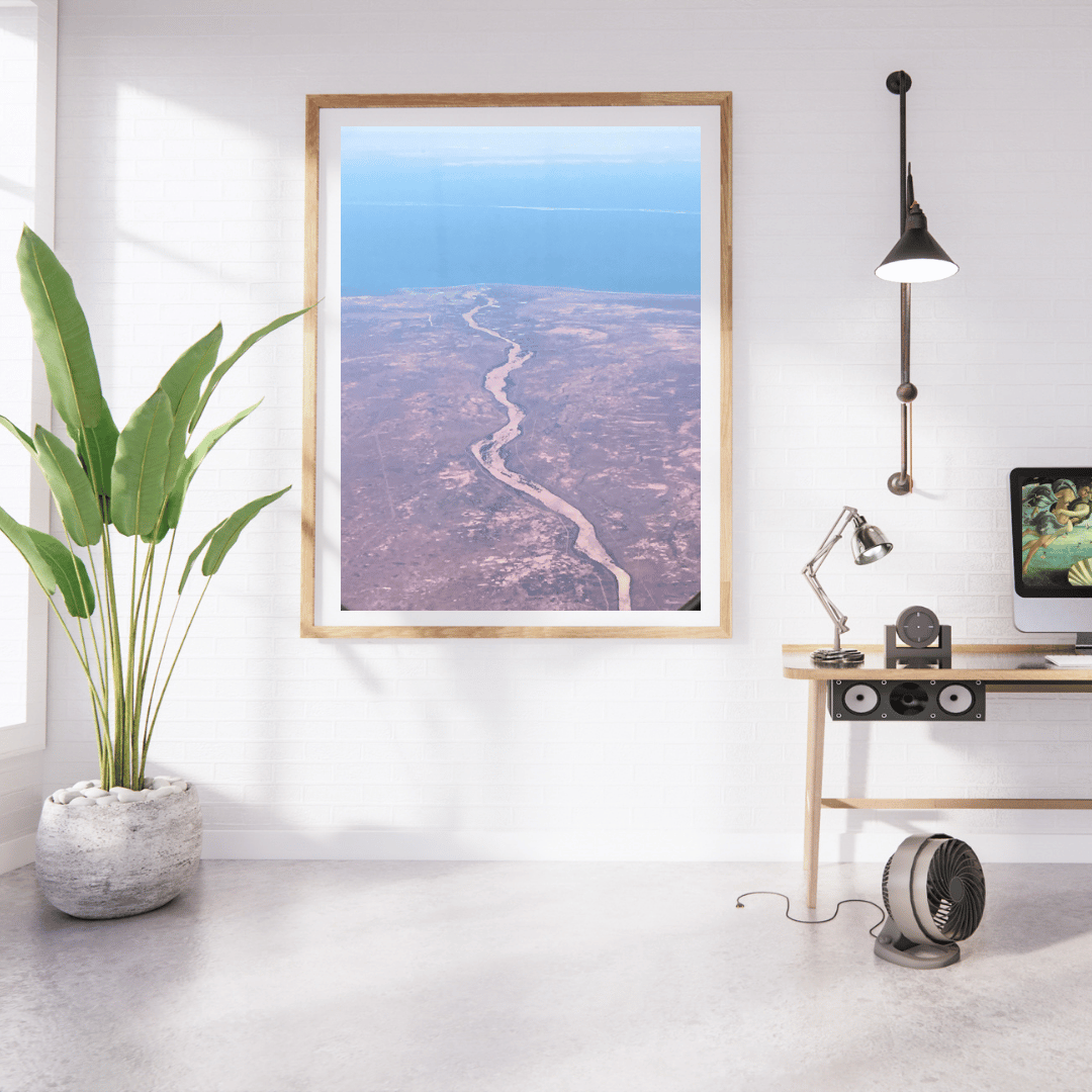 A framed vertical art print showing an aerial view of the crimson colour of the barren land in Australia, displayed on a white wall in a modern home office setting with a desk, lamp, and plant.