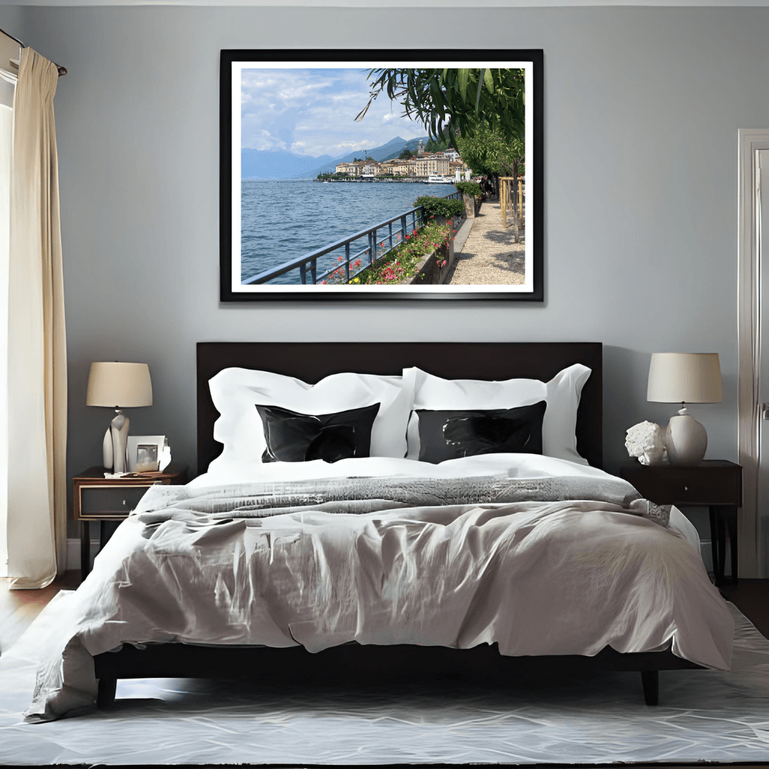 A framed landscape art print of a lakeside town hangs on a light gray wall above a bed in a modern, symmetrically styled bedroom.