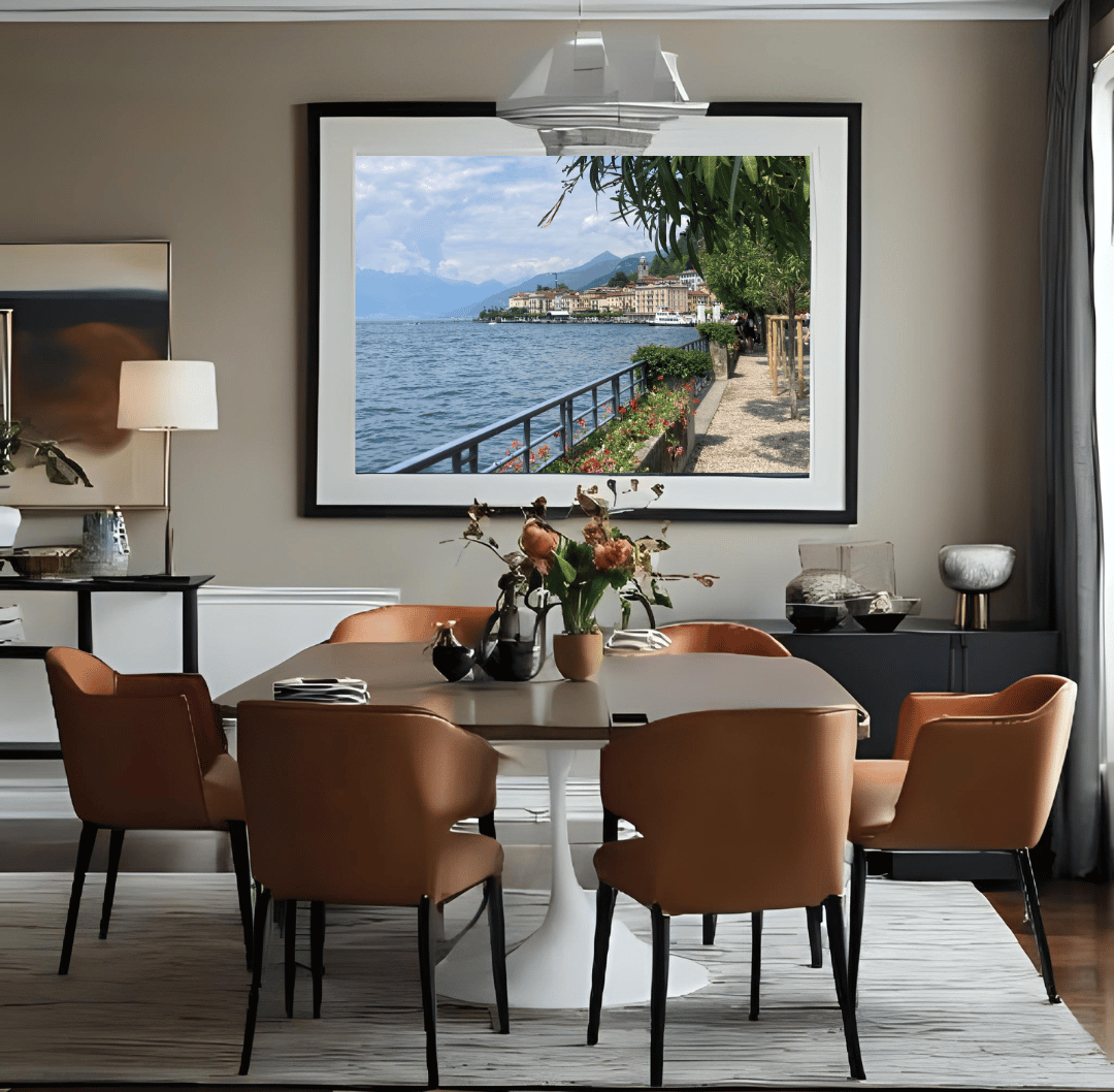 A large framed landscape photograph of a scenic  Lake Como and town hangs on a beige wall in a modern dining room setting with a wooden dining table and six brown upholstered chairs.
