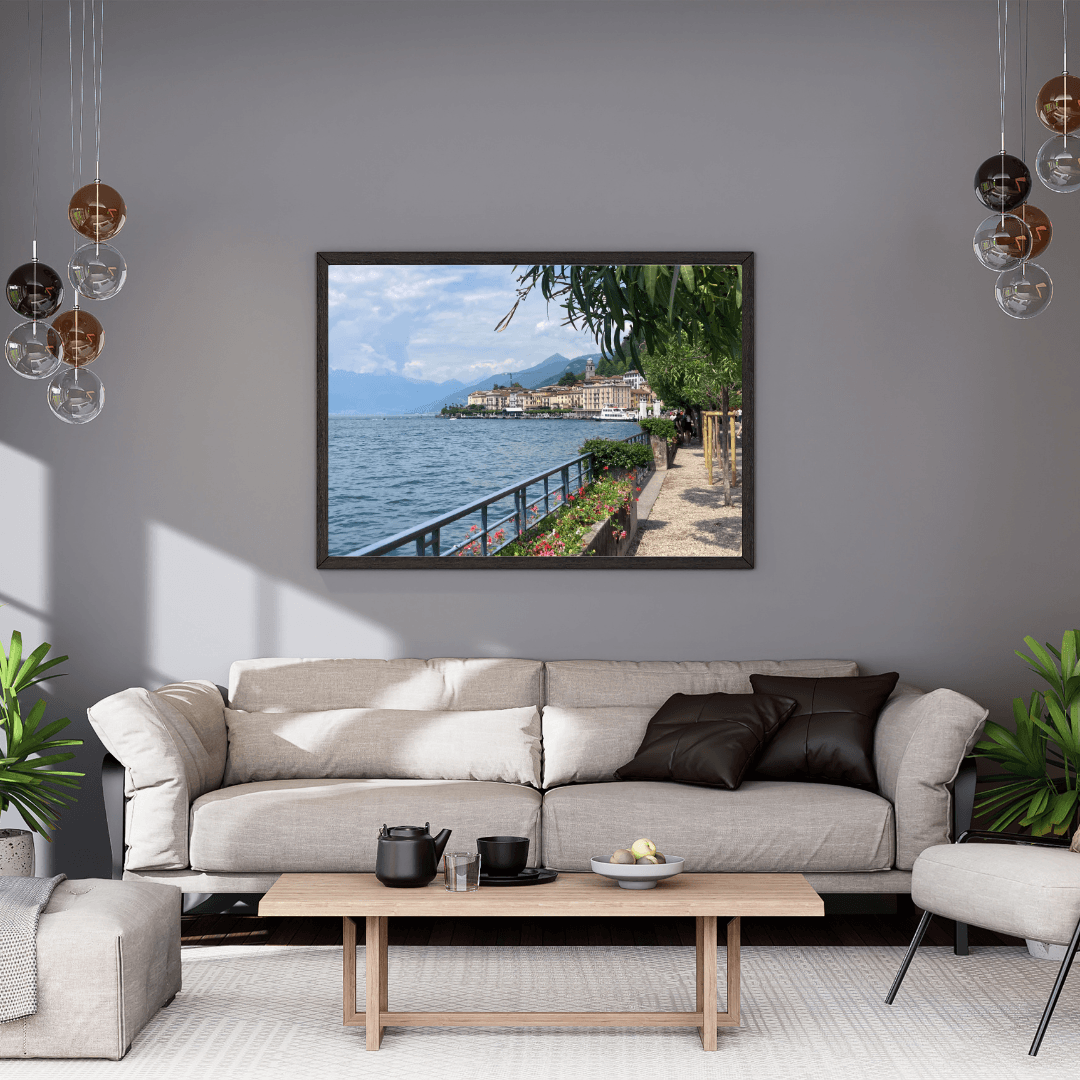 A framed landscape photograph of a scenic  Lake Como and town hangs on a gray wall above a modern beige sofa in a styled living room.
