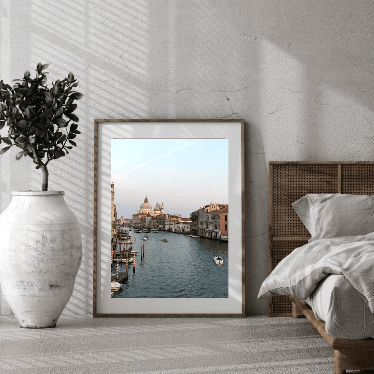 A framed art print depicting a scenic Venetian canal city view, styled in a modern bedroom setting next to a bed and a large white vase with a potted plant.