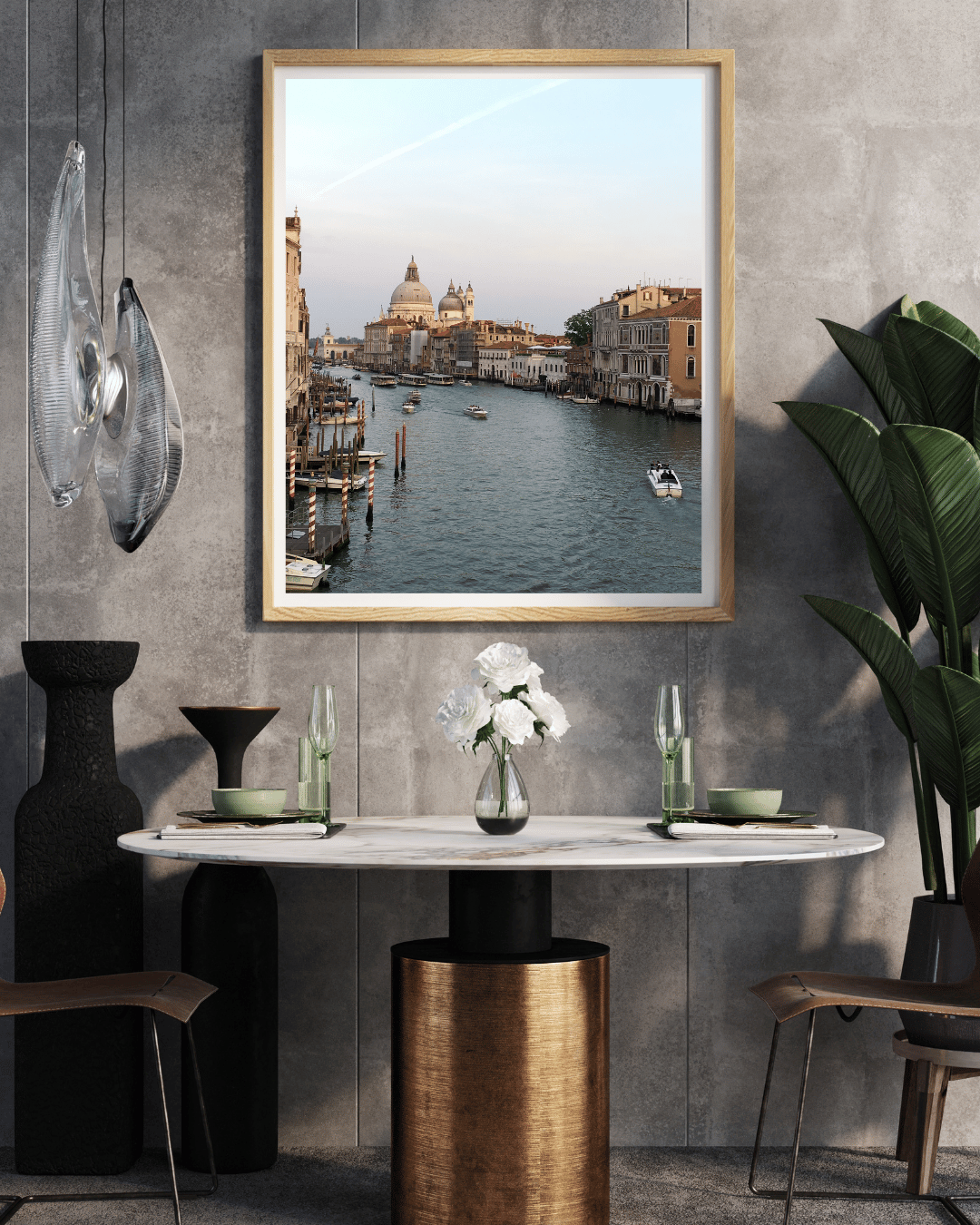 A framed art print depicting a scenic Venice canal with the St Mark's Cathedral in the background, styled on a gray concrete wall above a dining table in a modern interior setting.