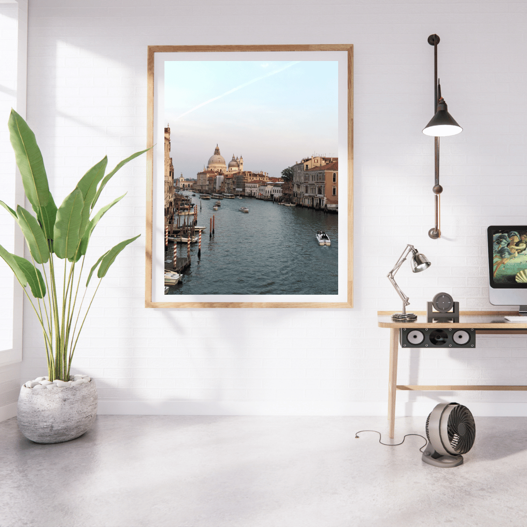 A framed art print of a scenic Venice canal with historic buildings and St Mark's cathedral, displayed on a white wall in a modern, sunlit room with a potted plant, desk, lamp, and computer monitor.