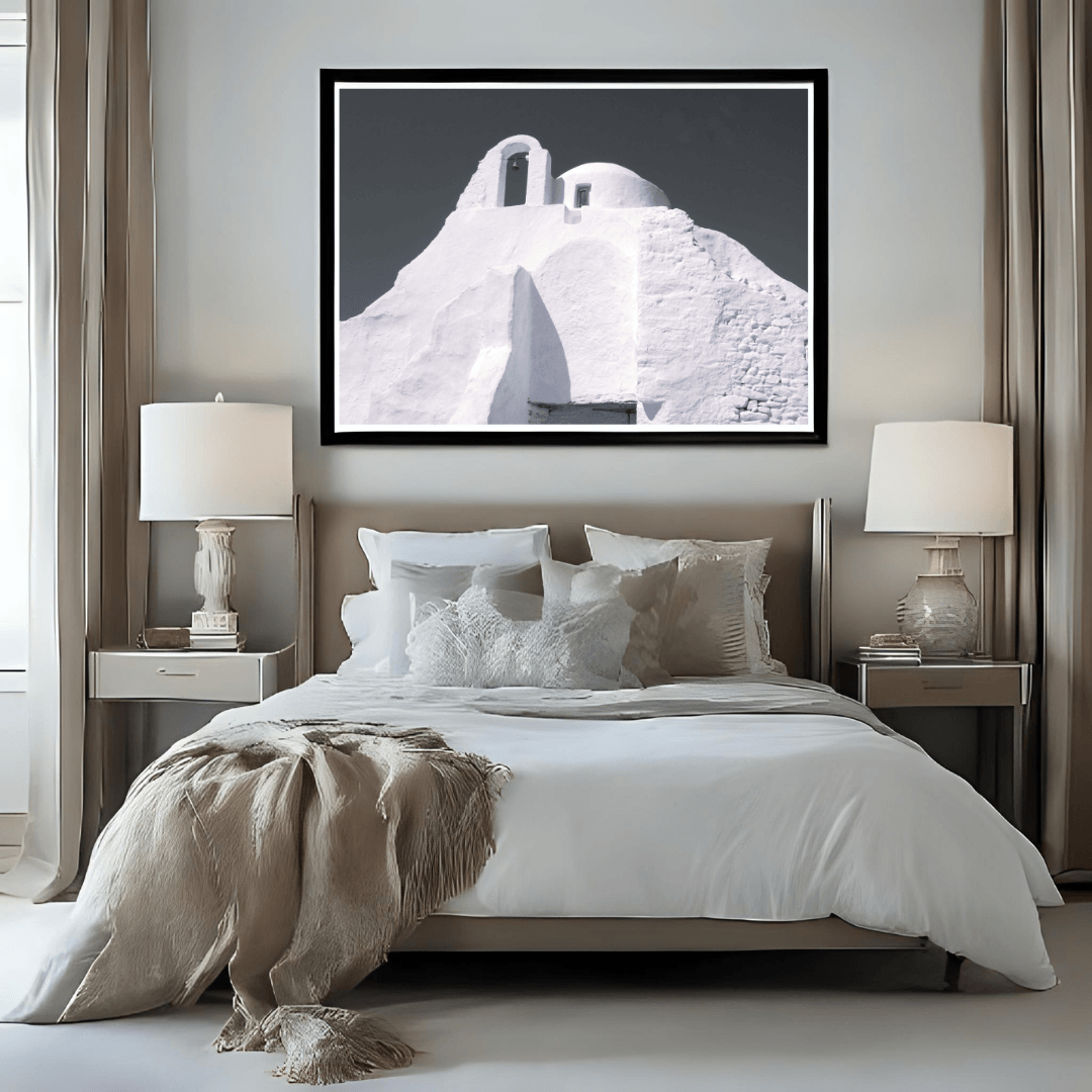 A framed black and white art print of a Santorini-style whitewashed buildings in Mykonos displayed on a neutral bed in a modern, symmetrical bedroom setting.