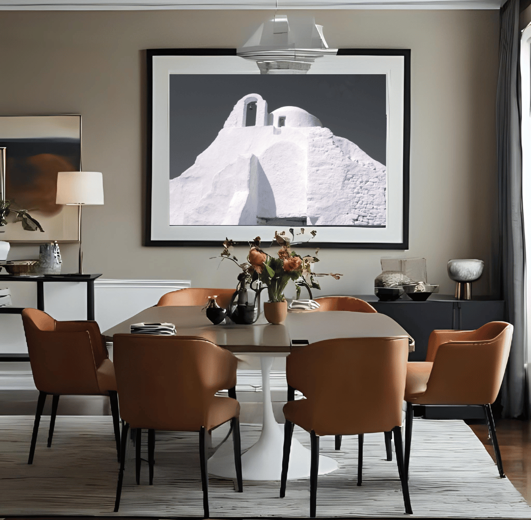 A styled dining room featuring a large black and white framed art print of a Santorini-style whitewashed buildings in Mykonos above a modern dining table and six brown upholstered chairs.
