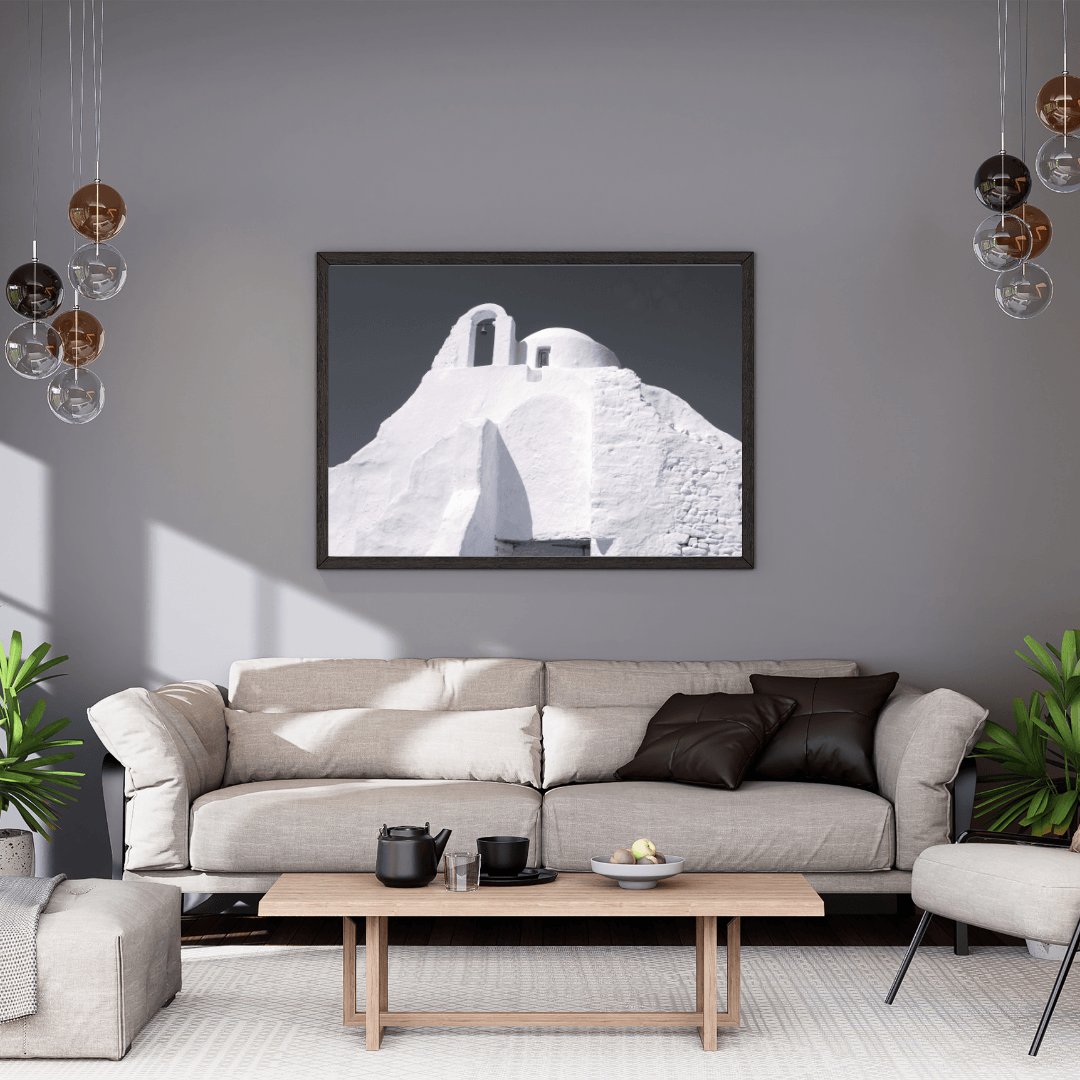 A framed black and white architectural art print of a Santorini-style whitewashed buildings in Mykonos displayed on a gray wall above a modern beige sofa in a styled living room.