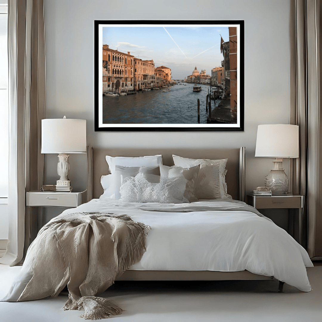 A framed art print of a Venice canal scene with historic buildings and a sunset sky, displayed on a light gray wall above a neatly made bed in a modern bedroom setting.