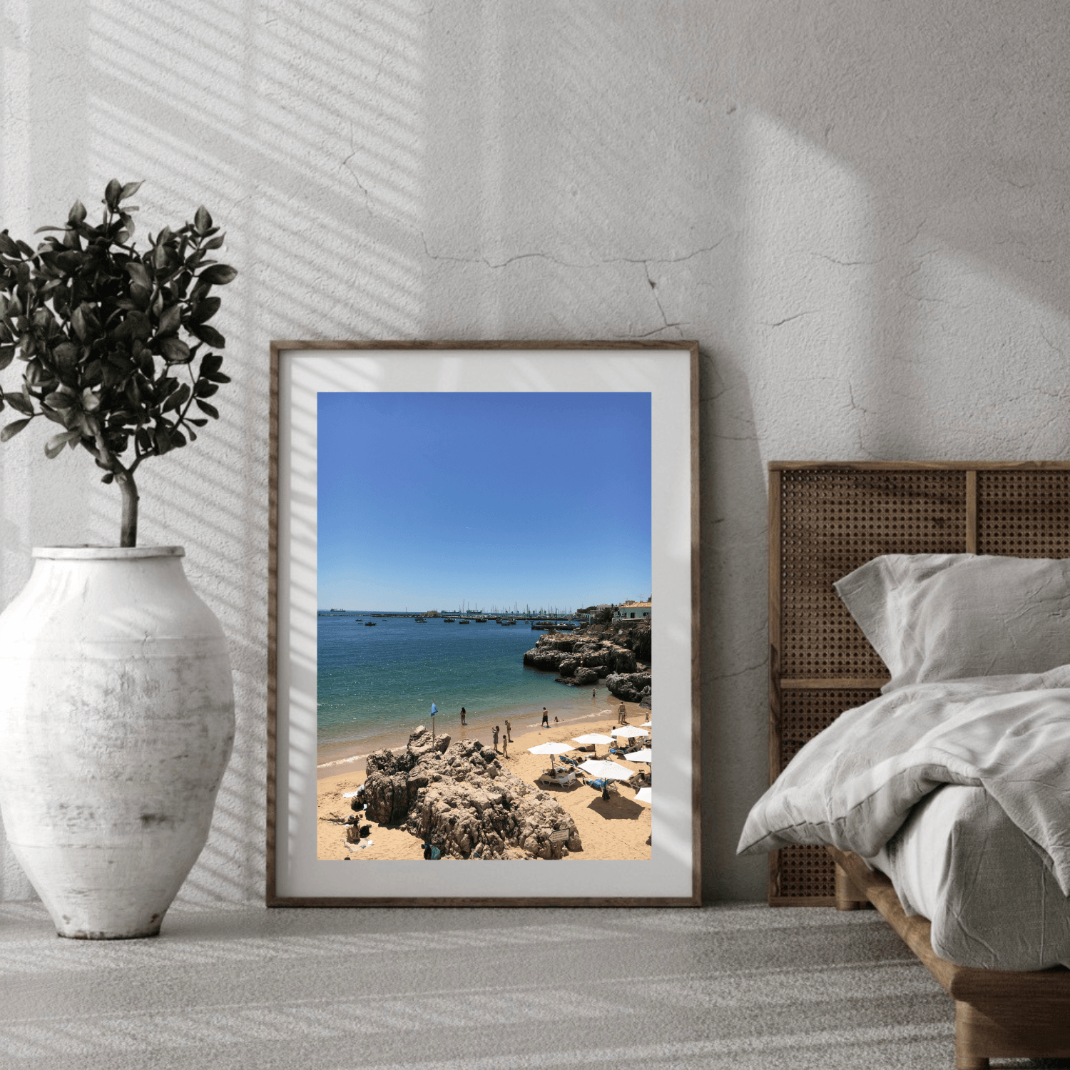A framed Portuguese beach landscape print leaning against a textured white wall in a styled bedroom, next to a bed and a white vase with a potted plant.