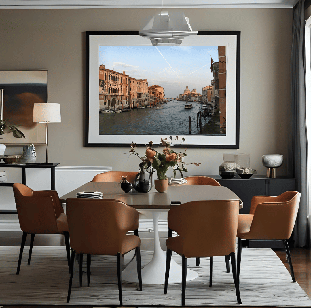 A large framed art print depicting a Venice canal scene hangs on a beige wall in a modern dining room setting with a dining table and six brown upholstered chairs.