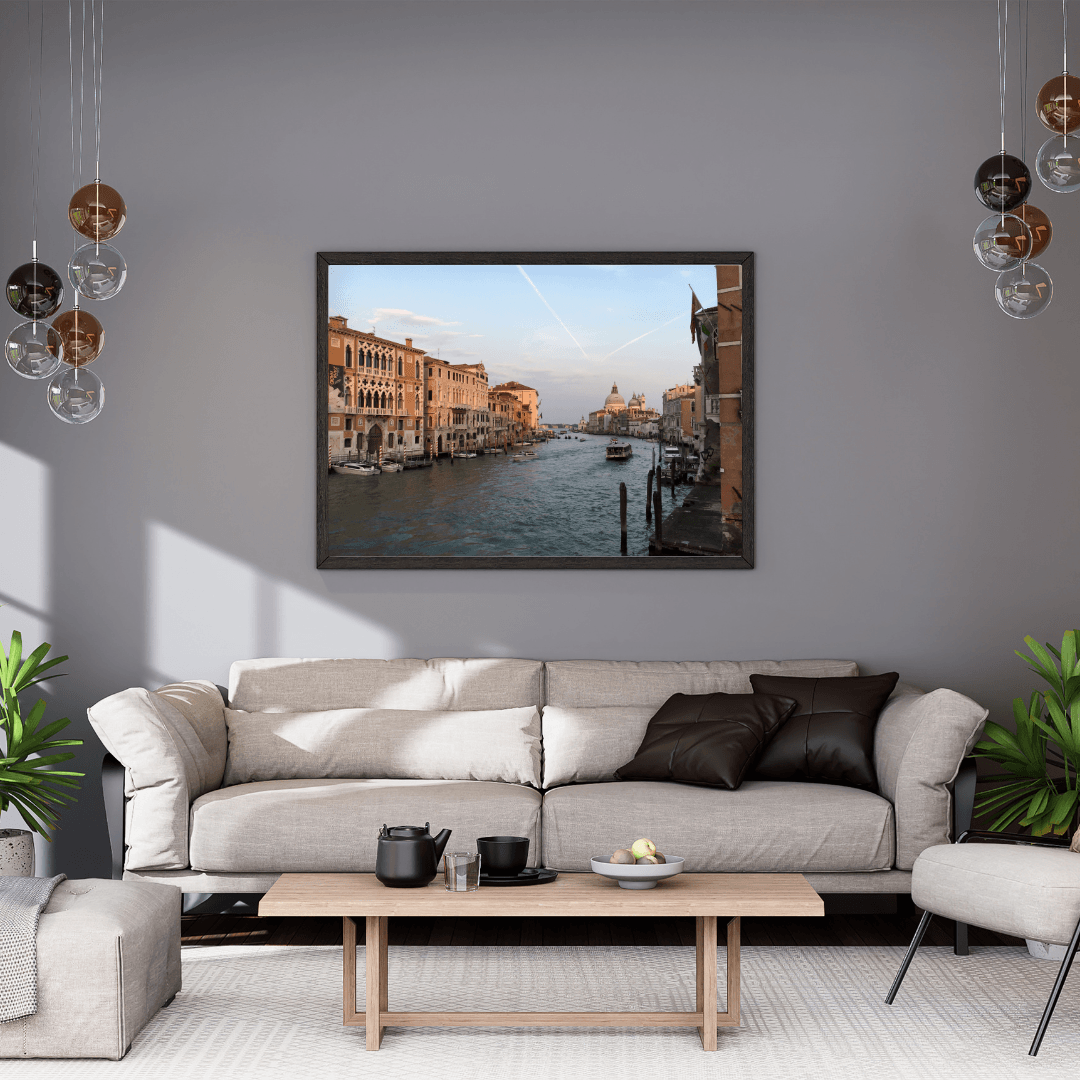 A framed art print depicting a scenic canal in Venice, displayed on a gray wall above a modern beige sofa in a styled living room.