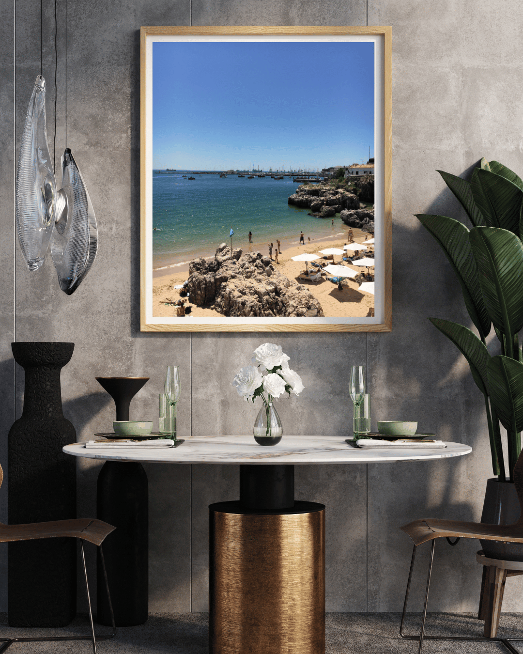 A framed beach print hangs on a textured gray wall above a dining table in a modern interior setting. The print shows a sunny coastal scene with blue water, sandy beach, rocks, and a small town under a clear blue sky, enclosed in a light wood frame.