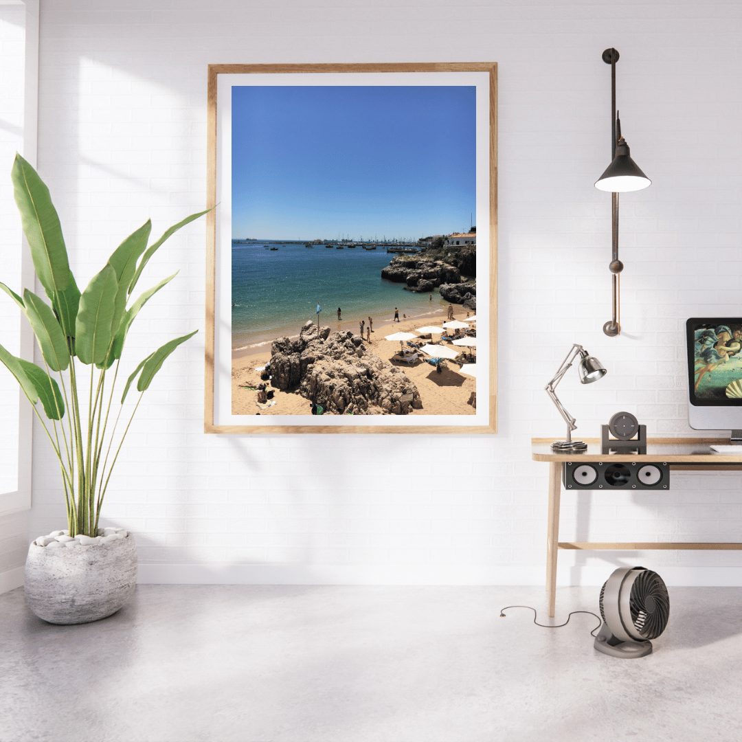 A framed beach print hangs on a white wall in a modern room setting with a potted plant, desk, lamp, and computer monitor visible.