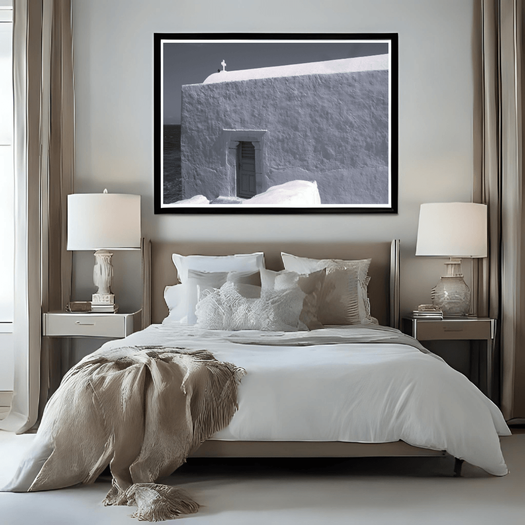 A framed black and white photograph of a small cross on top of a whitewashed building in Mykonos, Greece displayed as wall art above a neatly made bed in a modern, neutral-toned bedroom.