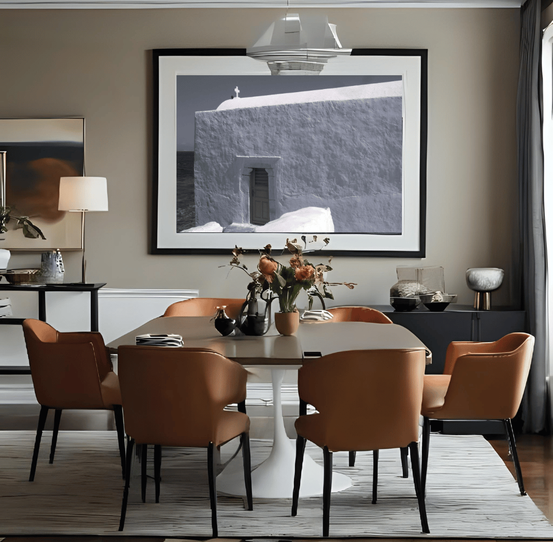 A styled dining room featuring a round white table surrounded by six tan leather chairs, set on a light gray rug beneath a large black-framed art print of a small cross on top of a whitewashed building in Mykonos, Greece.