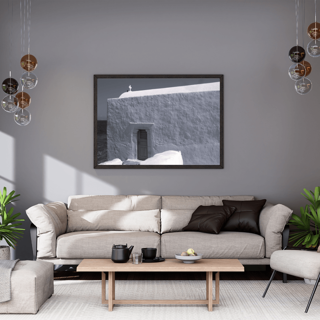 A framed black and white architectural photograph of a small cross on top of a whitewashed building in Mykonos, Greece hangs on a gray wall above a light beige sofa in a modern living room setting.