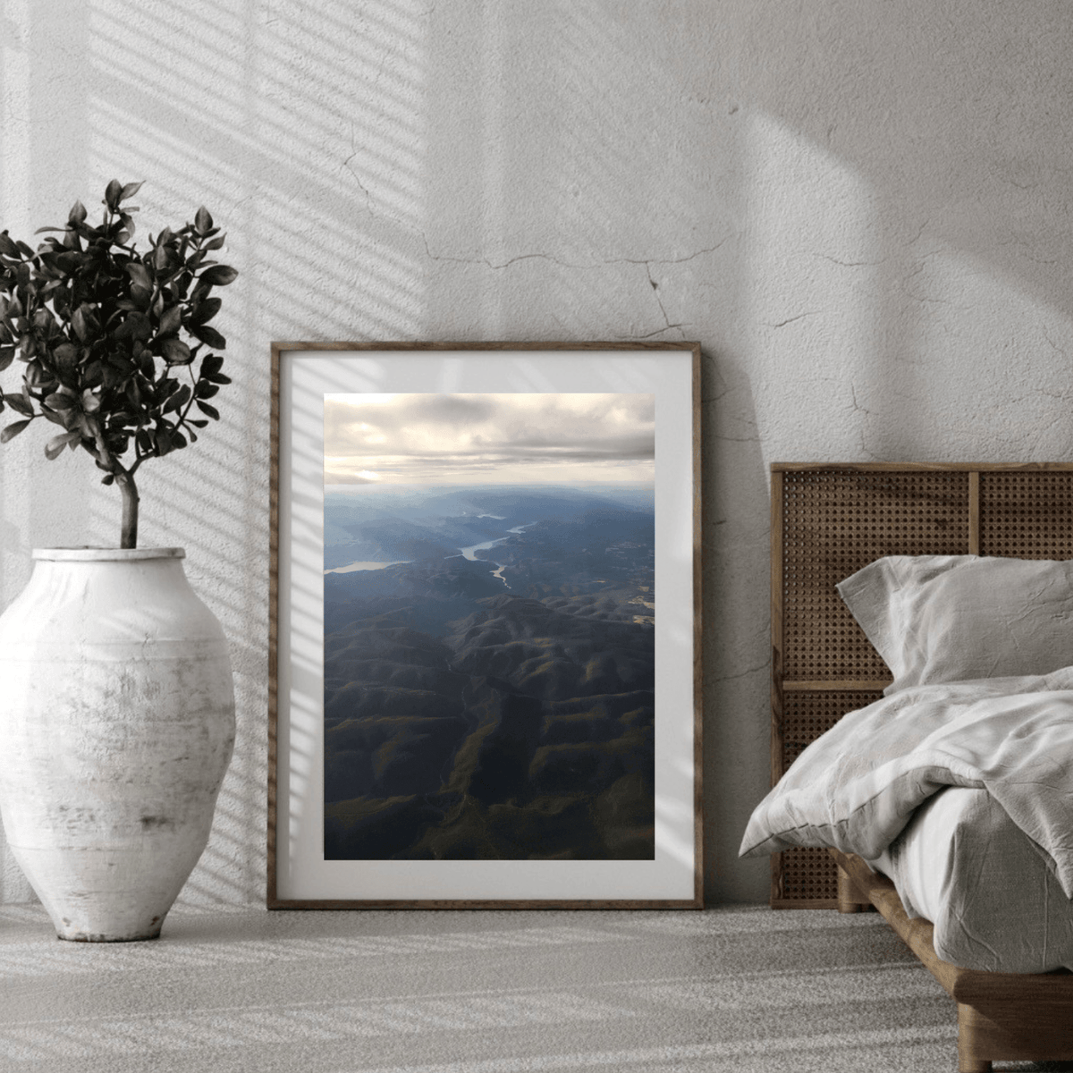 A framed landscape art print of an aerial view of the rays of light shining through clouds onto the dark blue and black mountains below, styled in a modern bedroom setting next to a bed and a white vase with a potted plant.