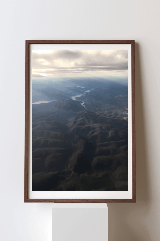 Heavens Above fine art travel photography print featuring an aerial view of the rays of light shining through clouds onto the dark blue and black mountains below in Australia
