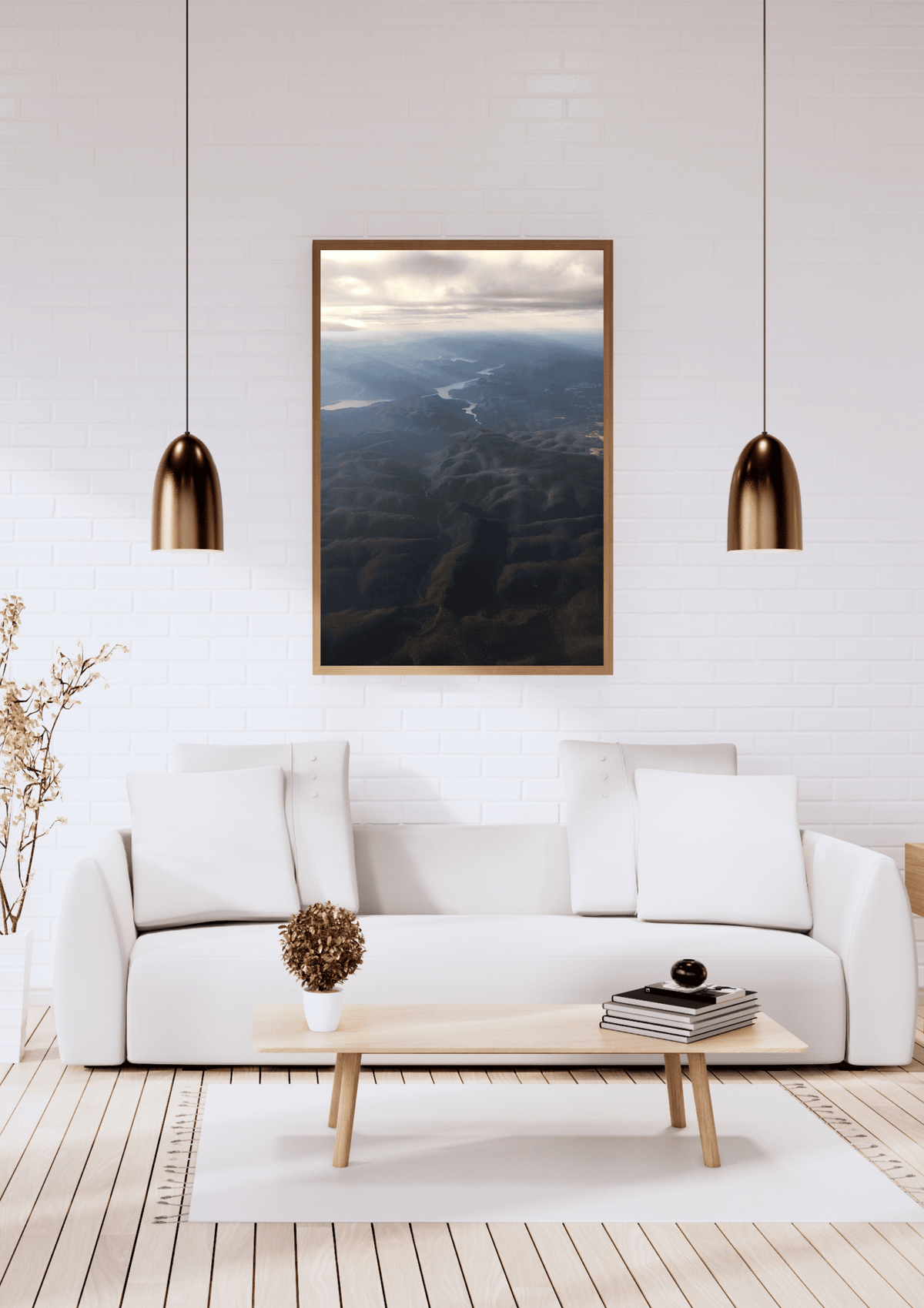 A framed landscape art print depicting an aerial view of the rays of light shining through clouds onto the dark blue and black mountains below, hung on a white brick wall above a modern white sofa in a bright living room.