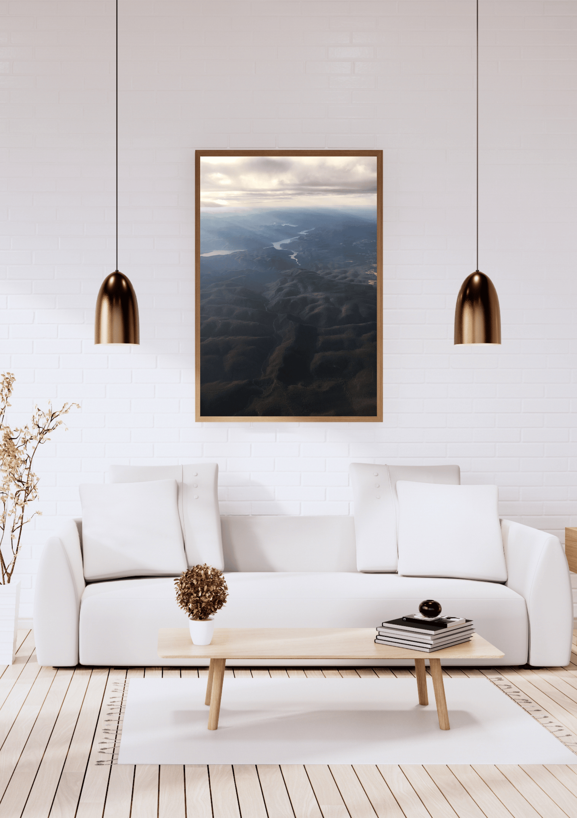 A framed landscape art print depicting an aerial view of the rays of light shining through clouds onto the dark blue and black mountains below, hung on a white brick wall above a modern white sofa in a bright living room.
