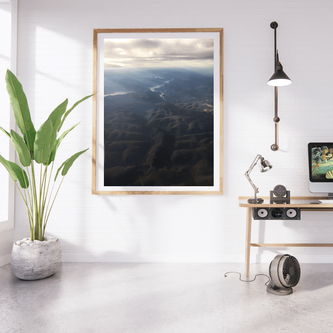 A framed vertical art print depicting an aerial view of the rays of light shining through clouds onto the dark blue and black mountains below, styled in a modern minimalist room with a potted plant, desk, lamp, and computer.