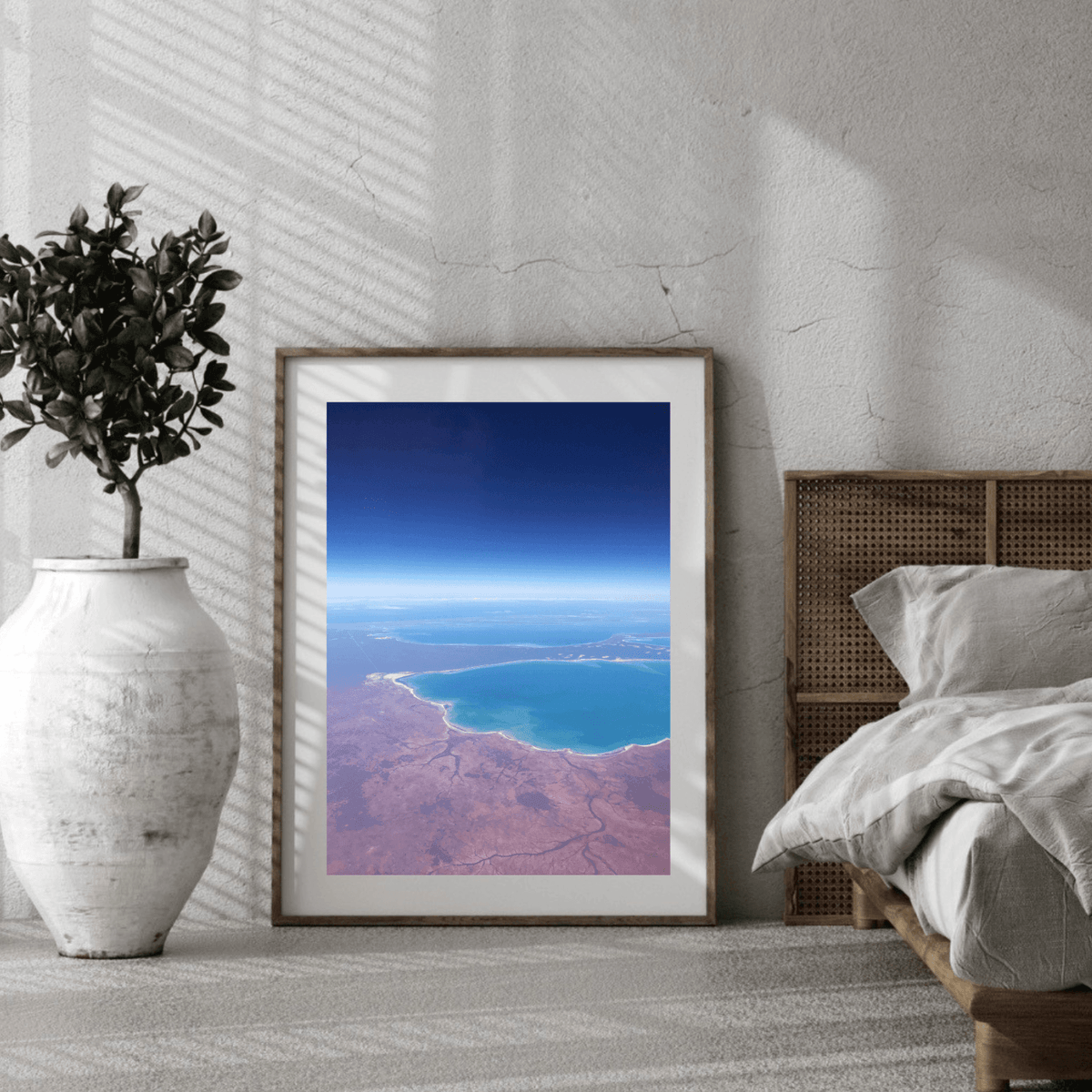 A framed aerial landscape art print of an aerial view of the deeply contrasting deep blue ocean against the red earth along the Western Australian coastline, styled in a modern bedroom setting next to a bed and a white vase with a plant.
