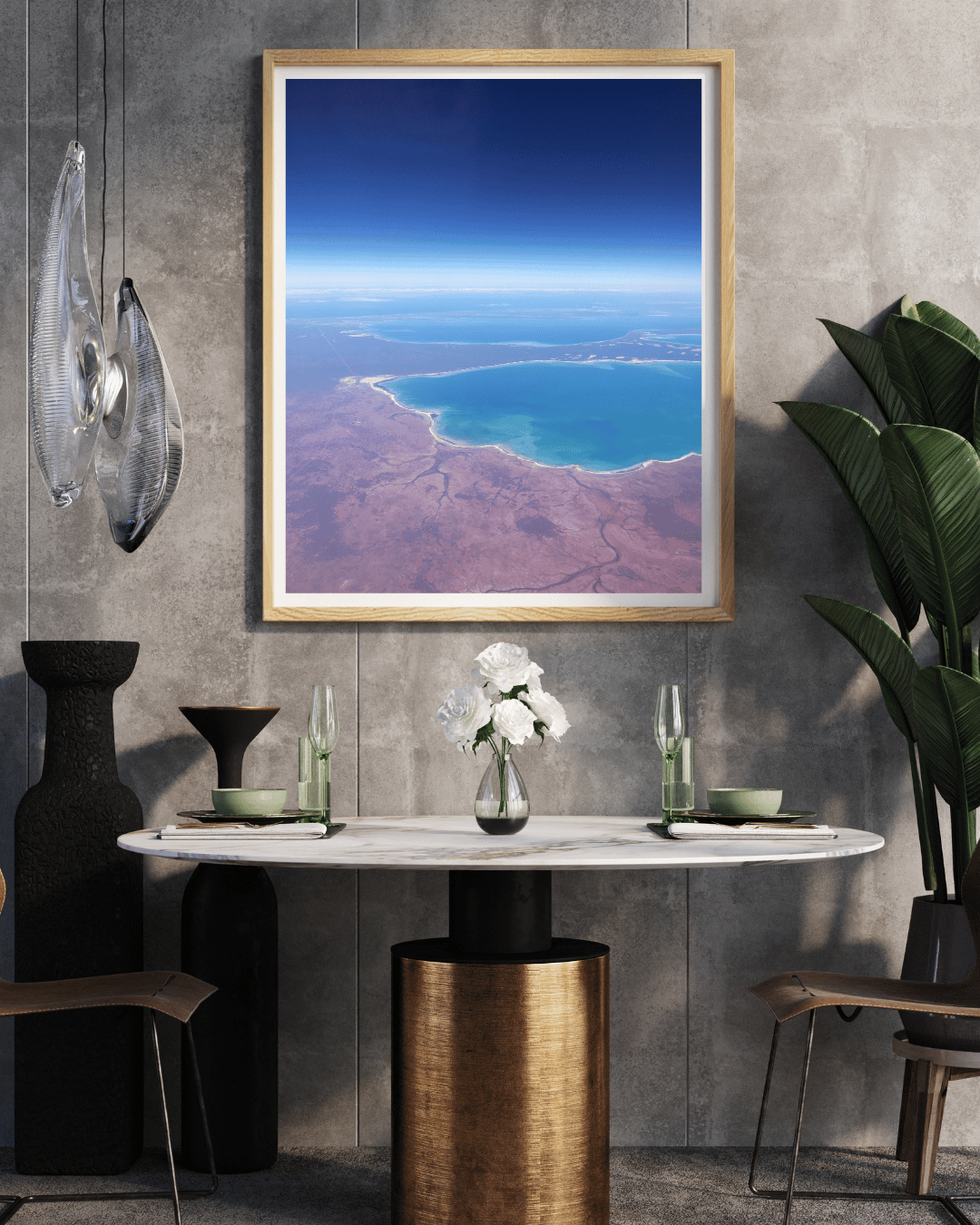 A framed aerial landscape photograph of an aerial view of the deeply contrasting deep blue ocean against the red earth along the Western Australian coastline, displayed on a gray wall above a dining table in a modern interior setting.