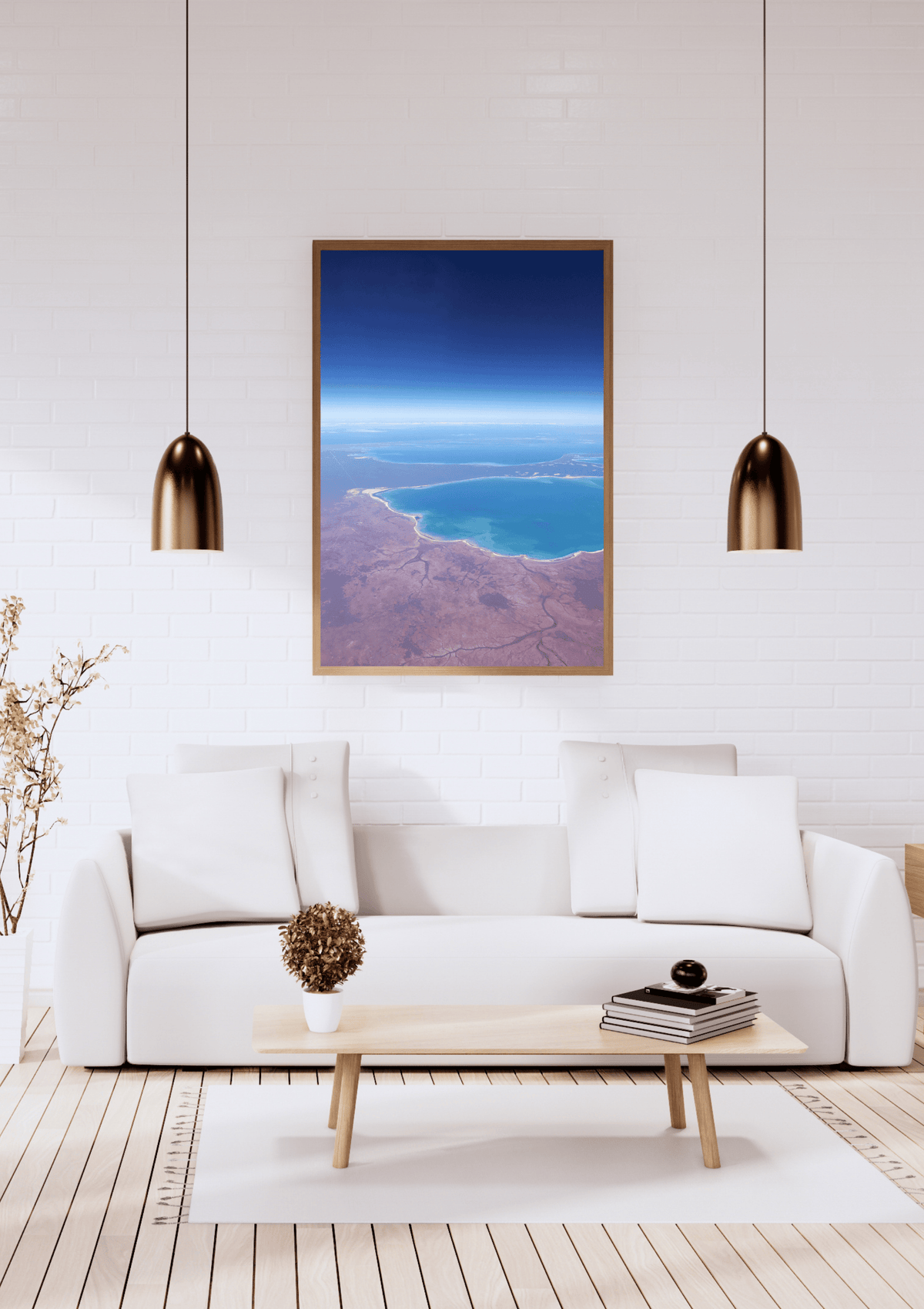 A framed art print depicting an aerial view of the deeply contrasting deep blue ocean against the red earth along the Western Australian coastline, displayed on a white brick wall above a modern white sofa in a bright living room.